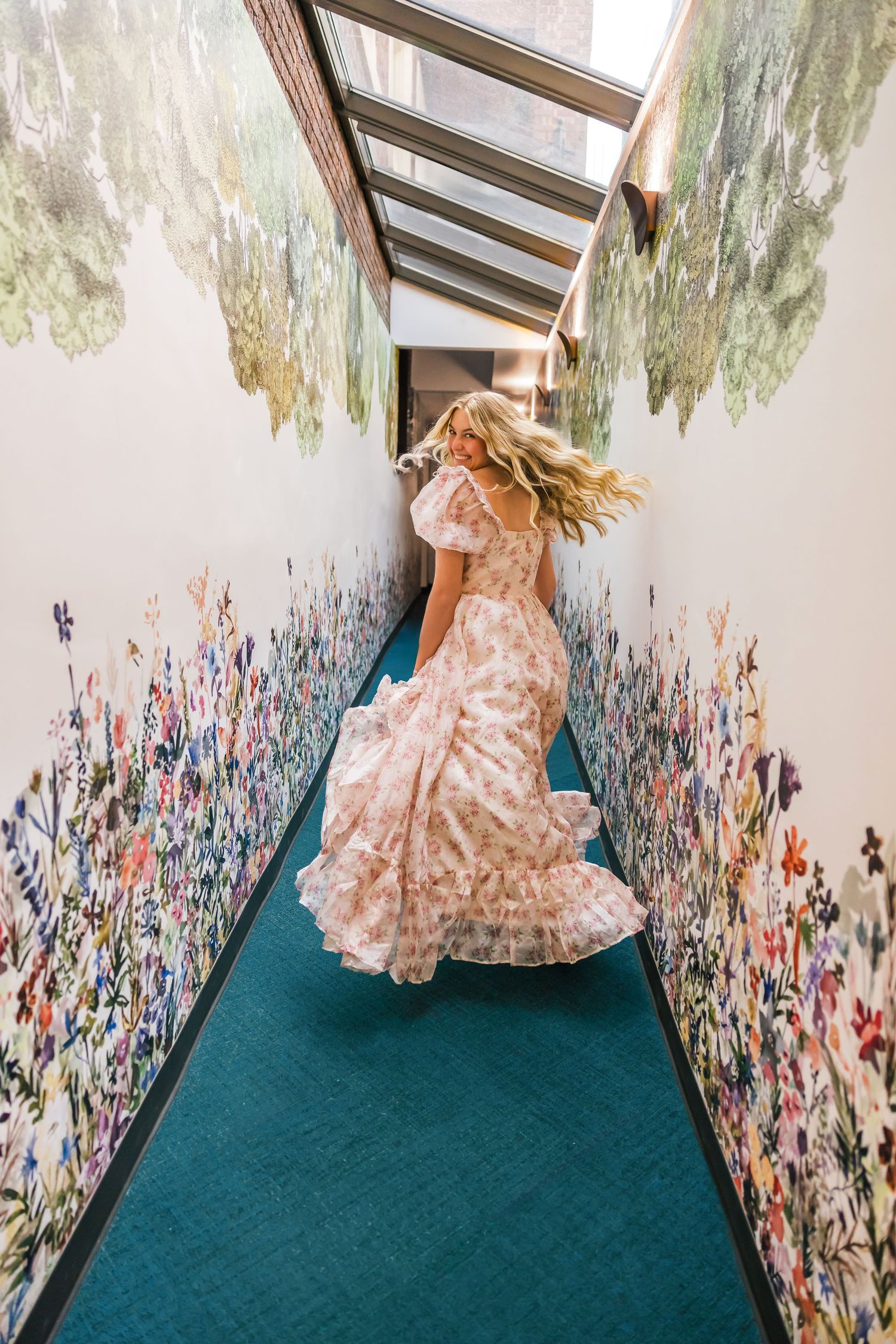 Person in flowing pink dress spins in hallway with floral wallpaper and skylight.