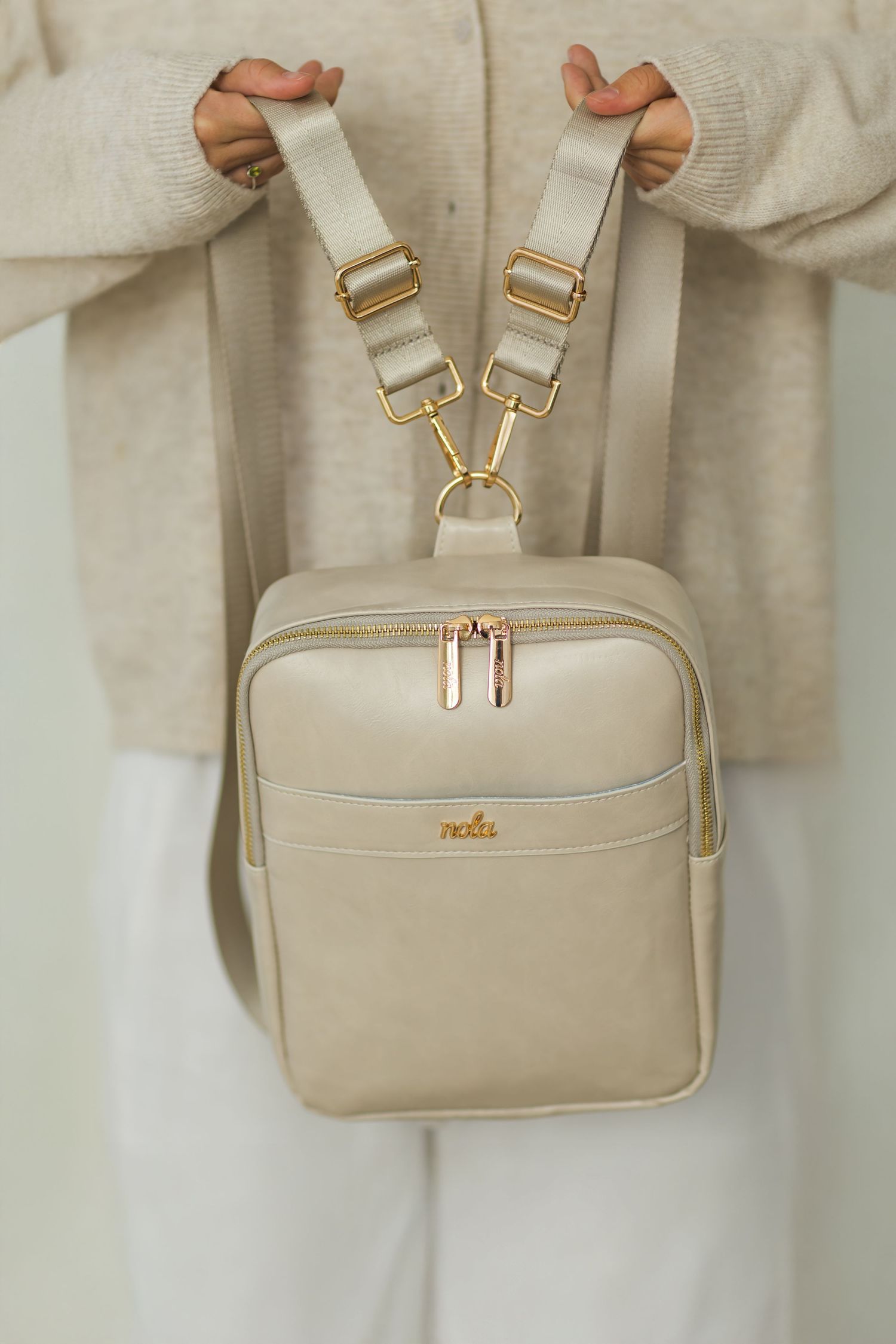 Cream colored leather backpack with adjustable straps being held up against neutral background.