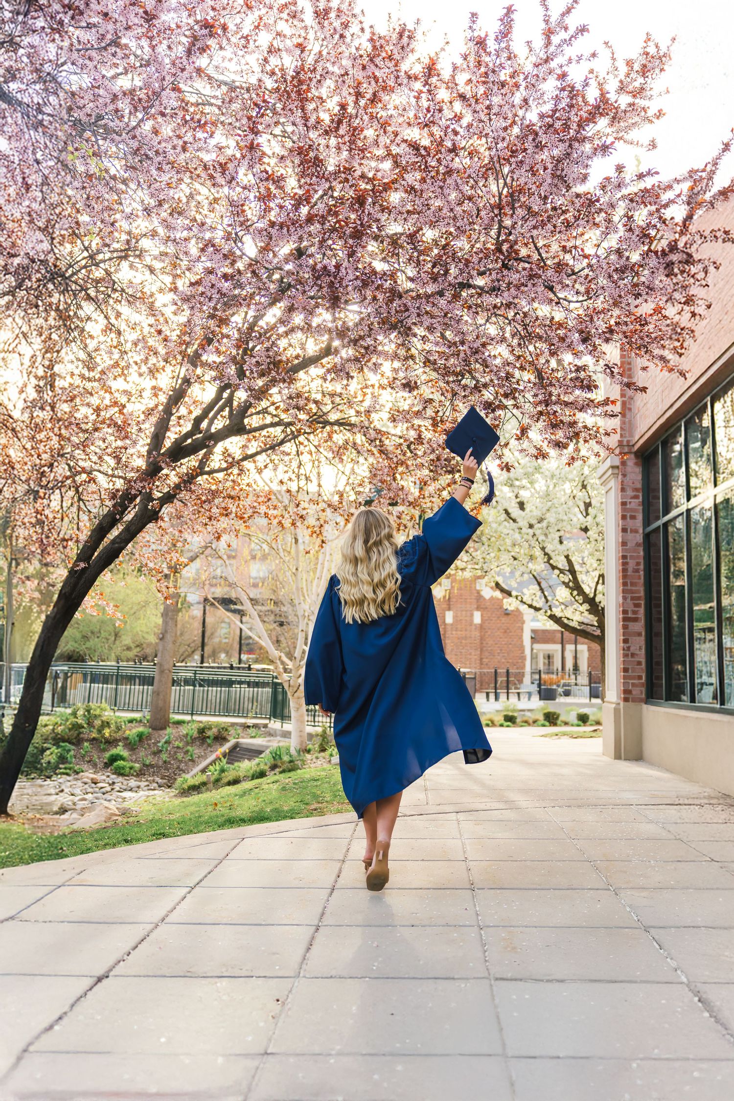 Graduate in blue cap and gown jumps joyfully under blooming cherry blossom trees.
