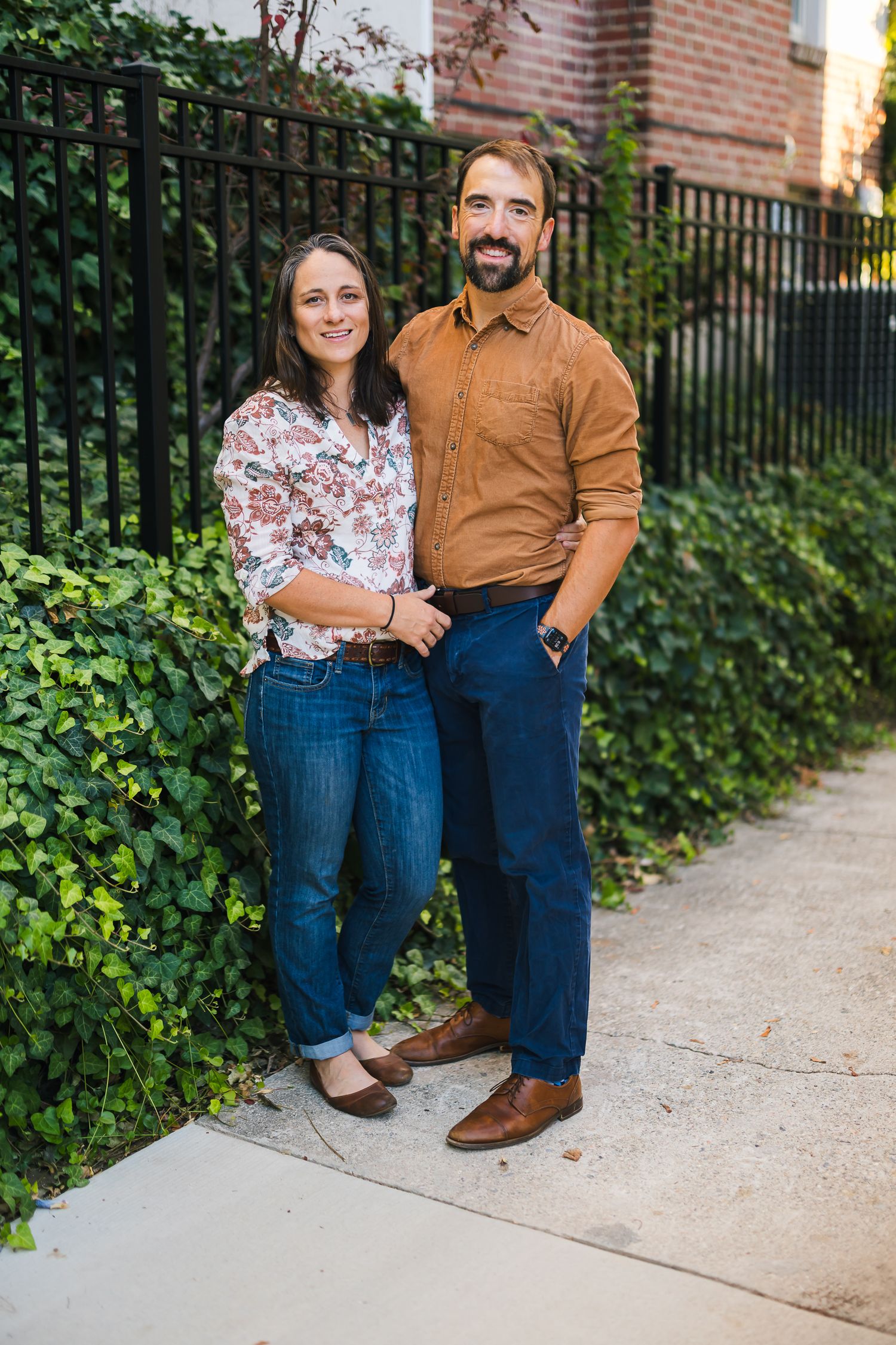 Couple in casual attire poses together on concrete path with garden and iron fence background.