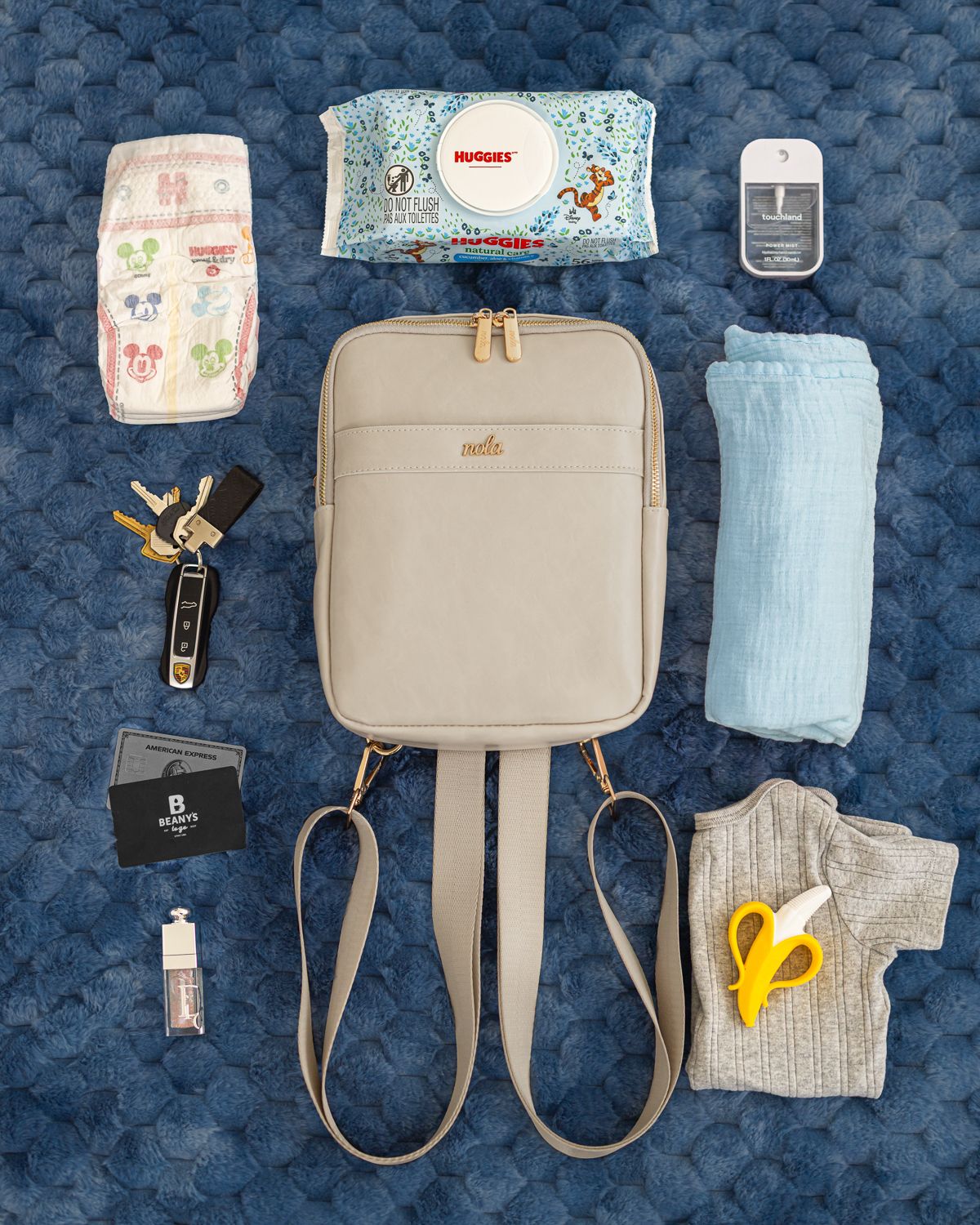 Flatlay of diaper bag contents including wipes, diapers, bottles, and changing pad arranged on blue textured background.
