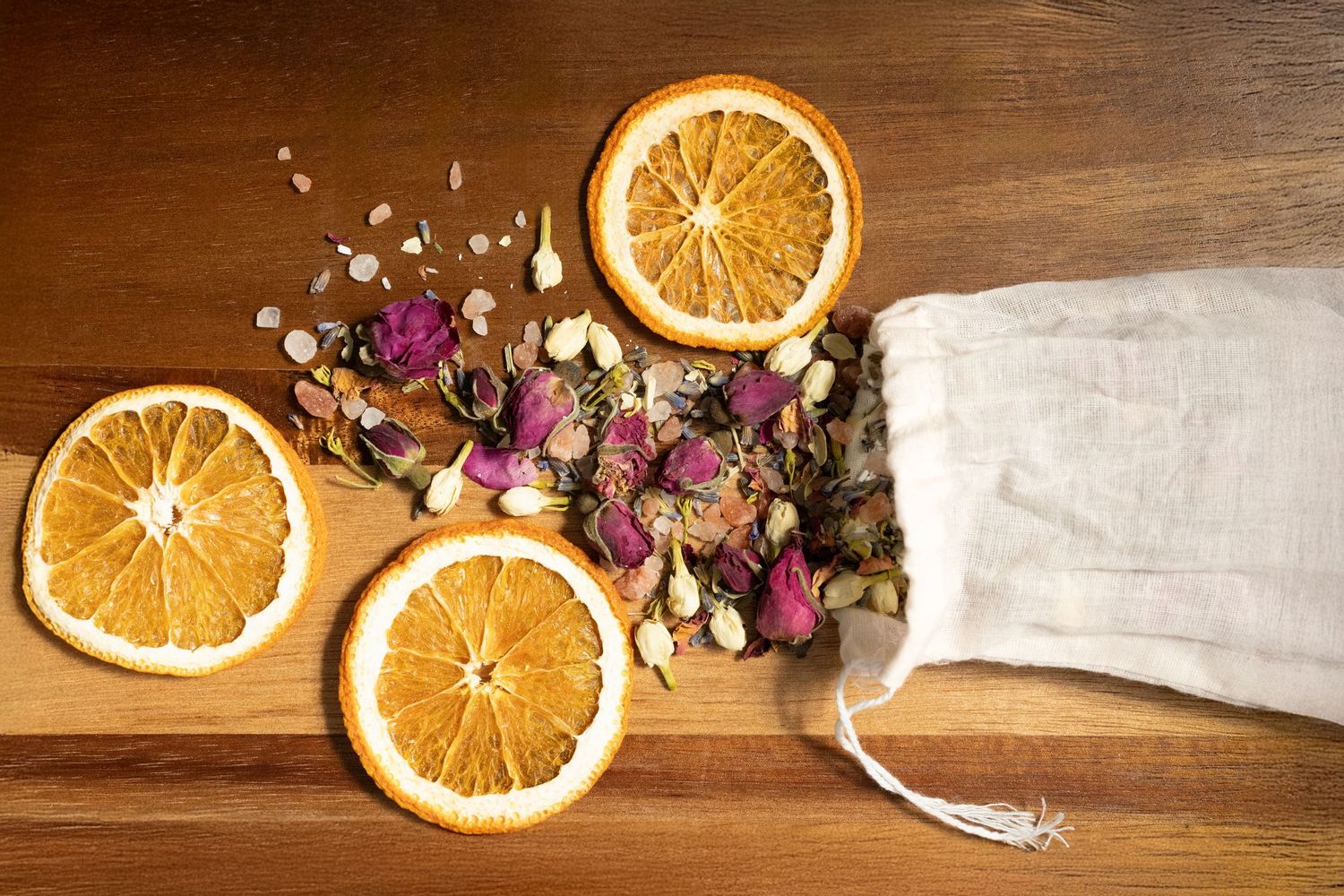 Dried orange slices and dried purple flower petals spill out of a white linen sachet on a wooden surface.