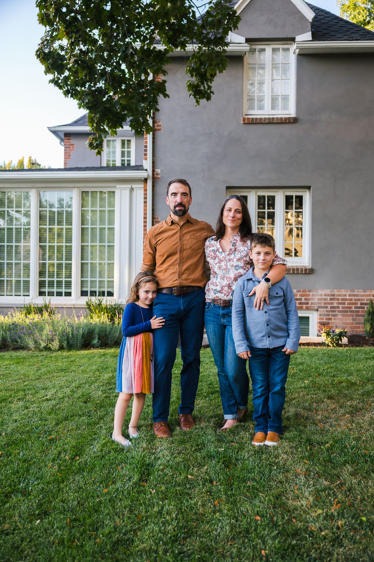 A family of four poses in front of their grey house with white trim windows.