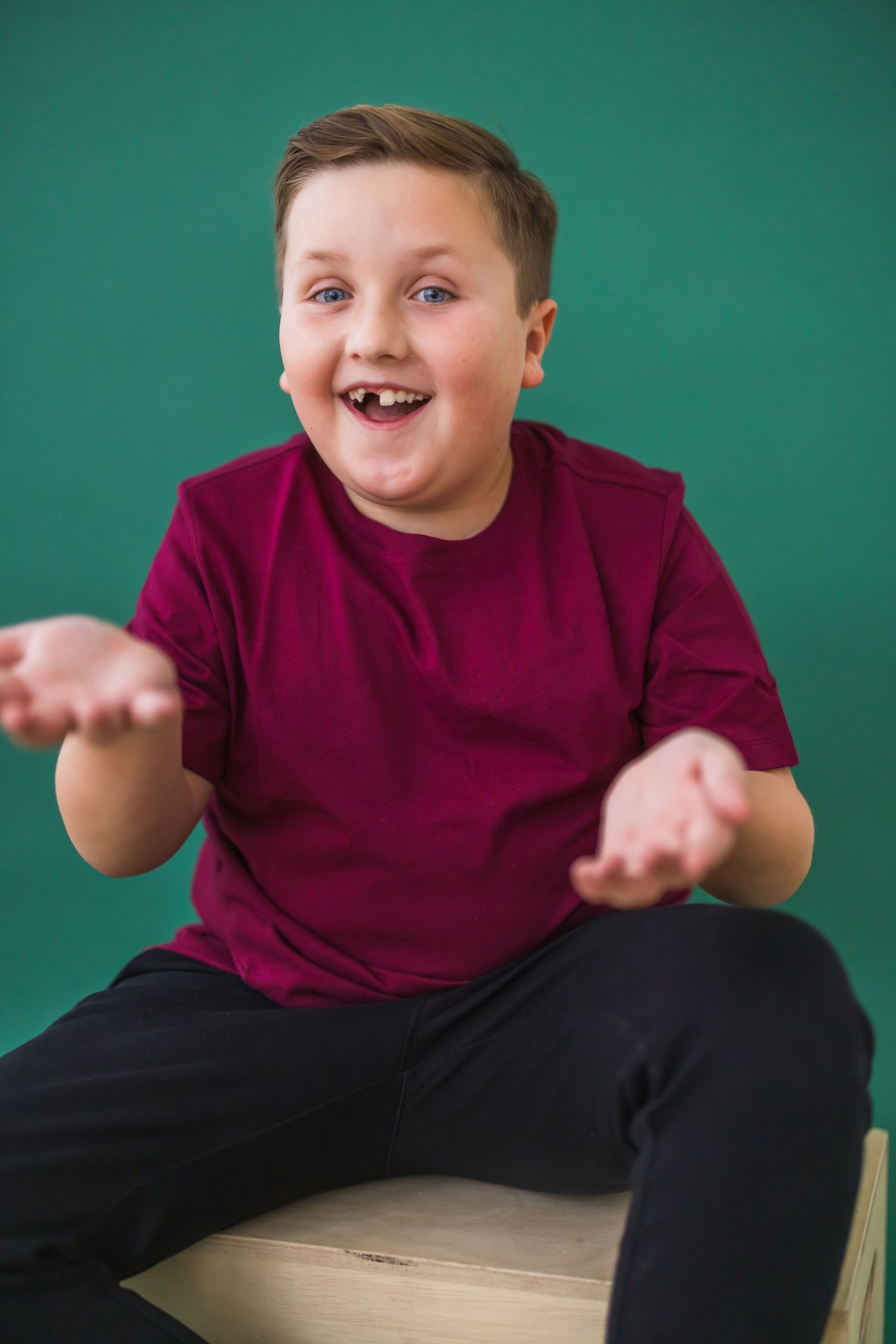 A child in a burgundy shirt and black pants sits on a surface making expressive gestures against a teal background.