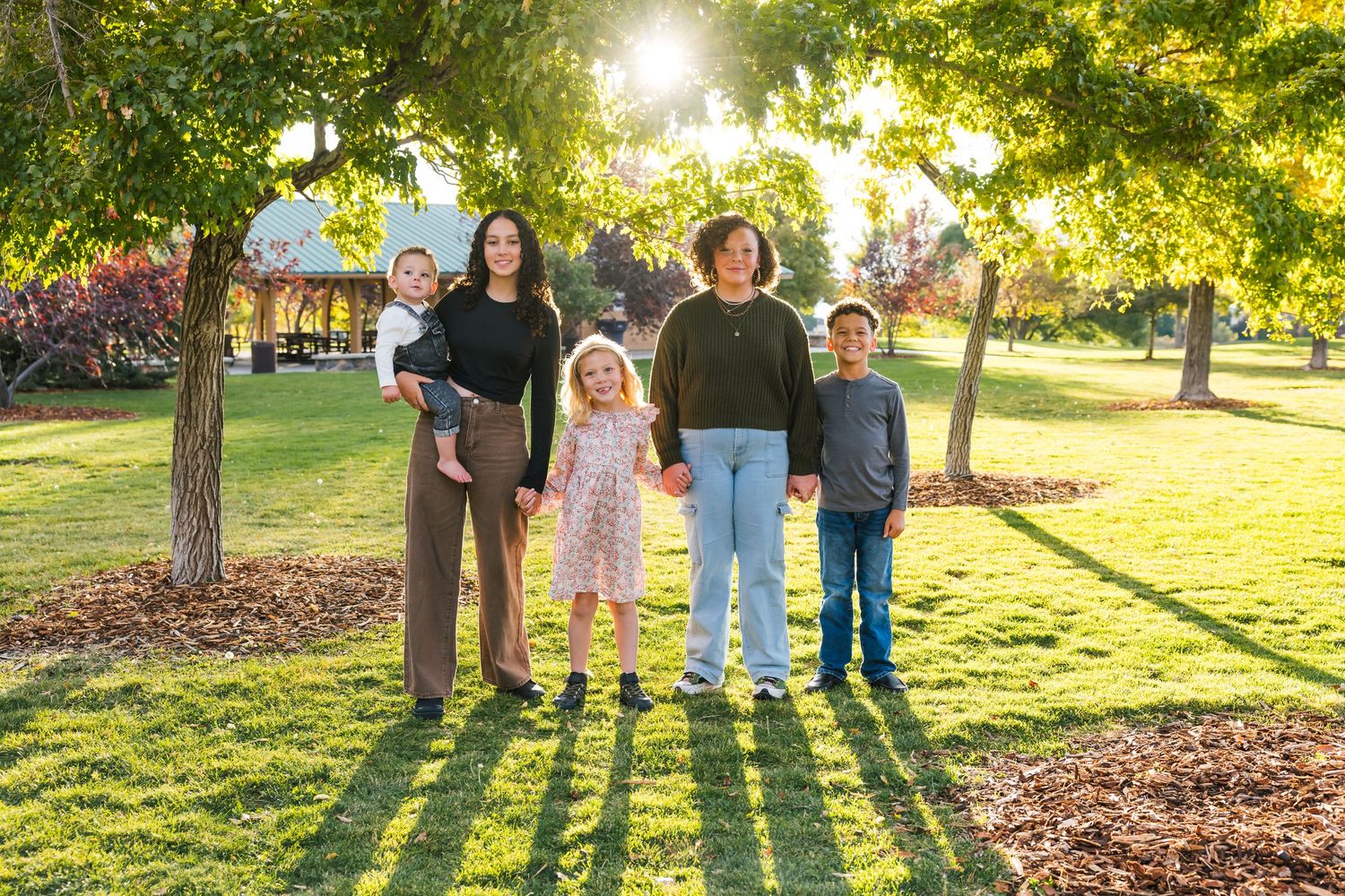 A family of five stand together in a sunlit park during golden hour with trees and mulch patches in the background.