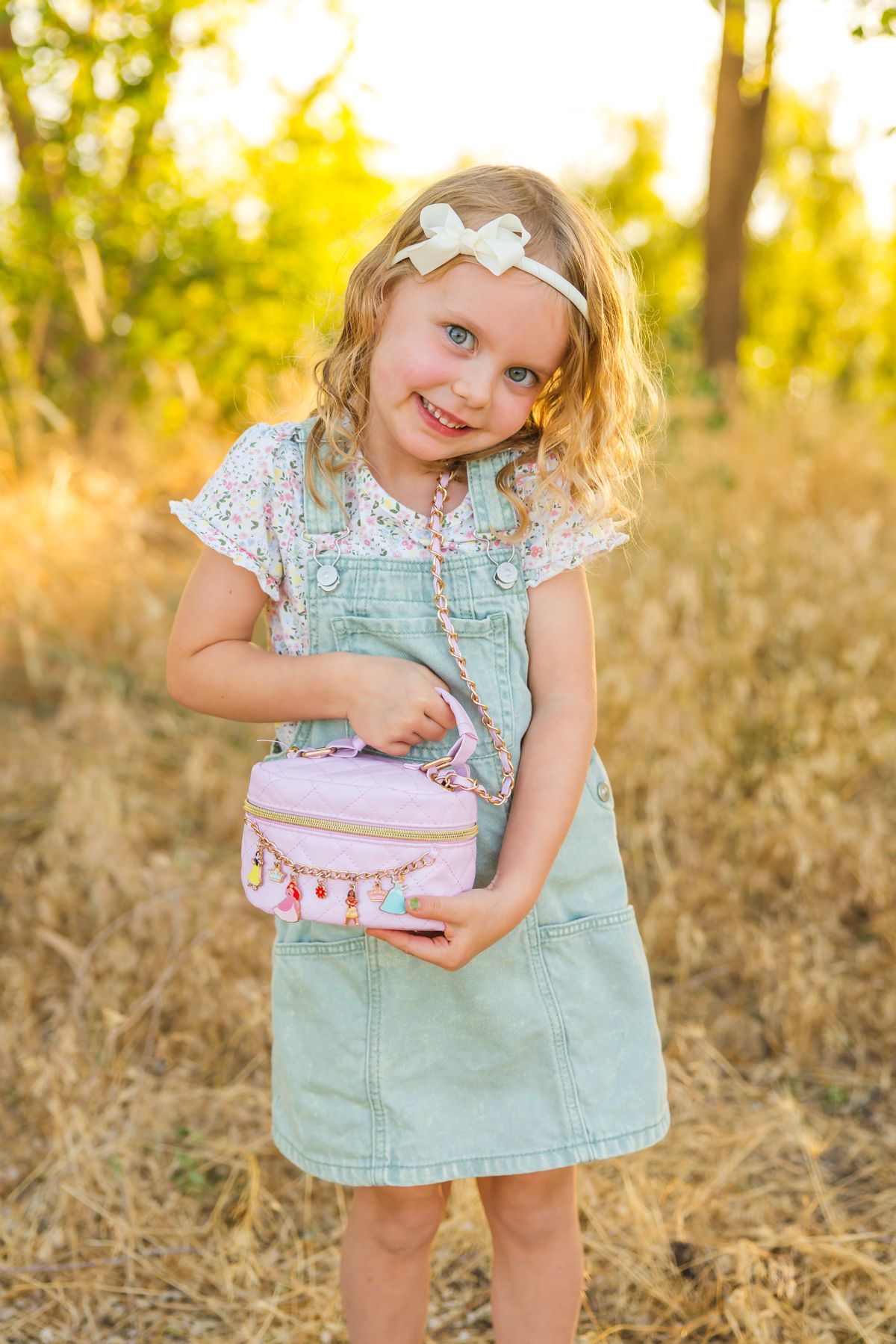 Child in mint dress holds small pink box while posing in sunlit outdoor setting.