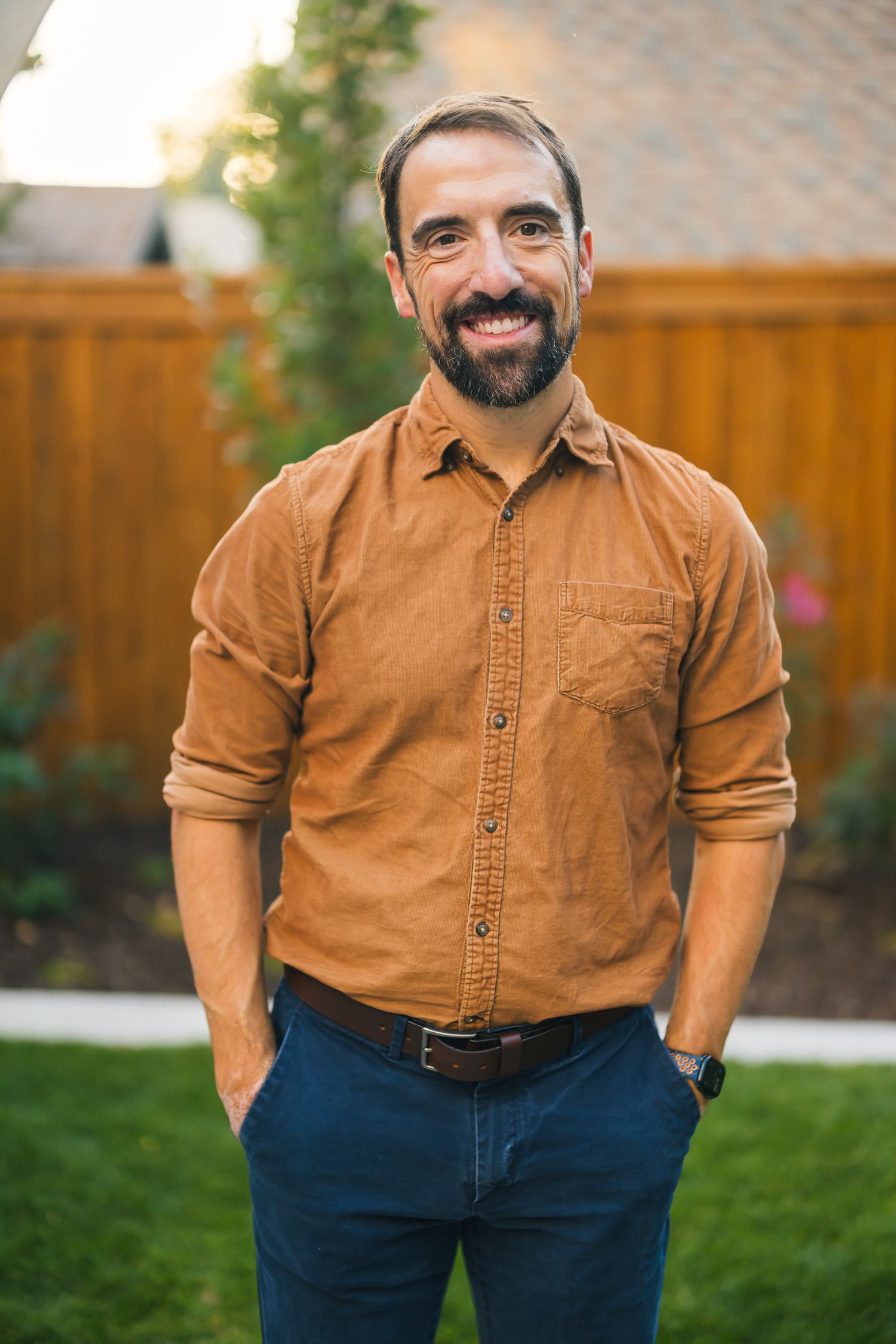 Professional portrait of person in brown button down shirt and navy pants standing in backyard garden setting.