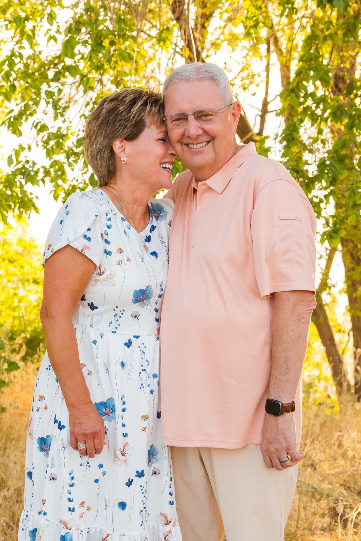 A couple wearing summer outfits shares a tender moment outdoors with golden sunlit trees in the background.