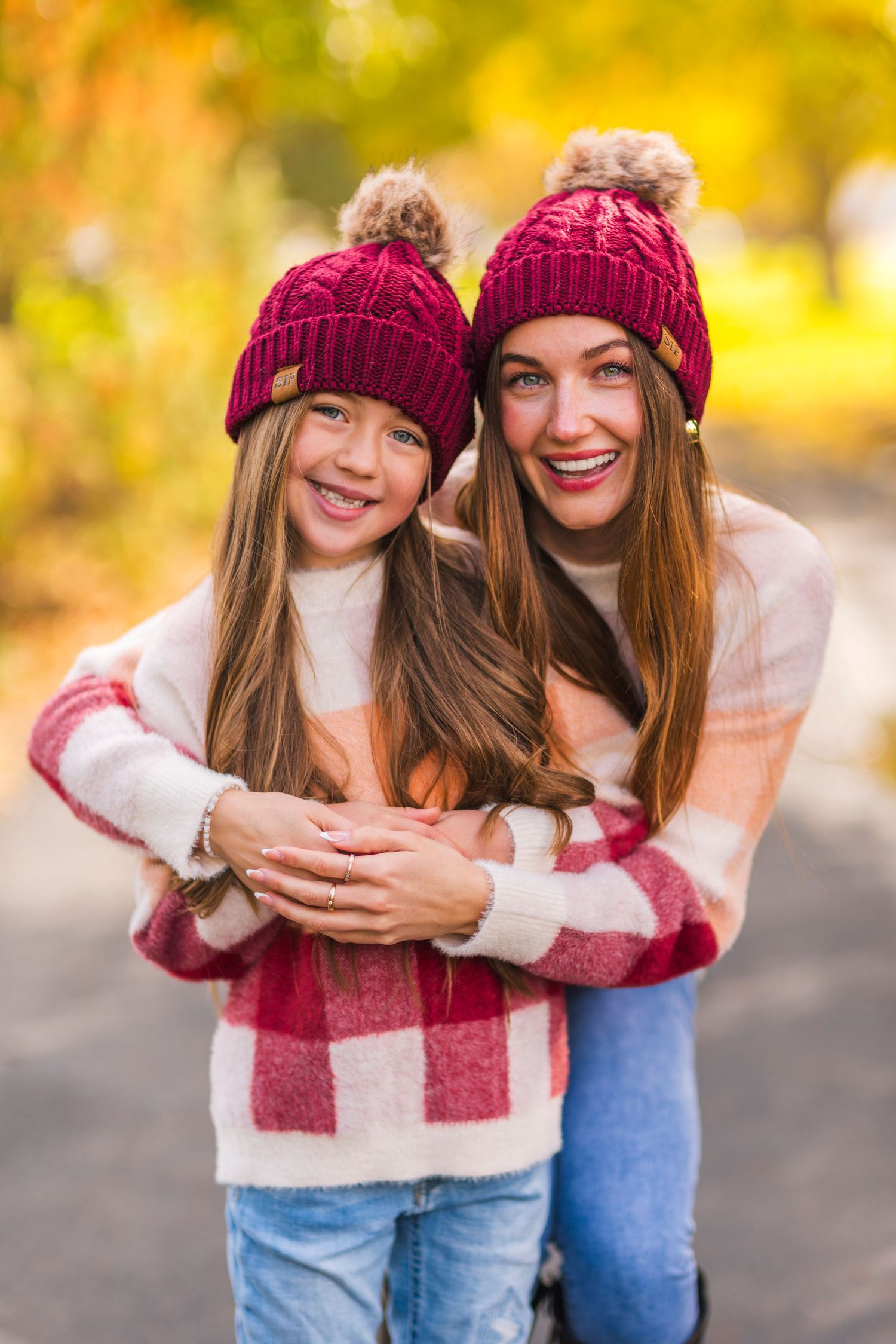 Two people wearing matching burgundy knit beanies and plaid sweaters embrace while smiling outdoors in autumn.