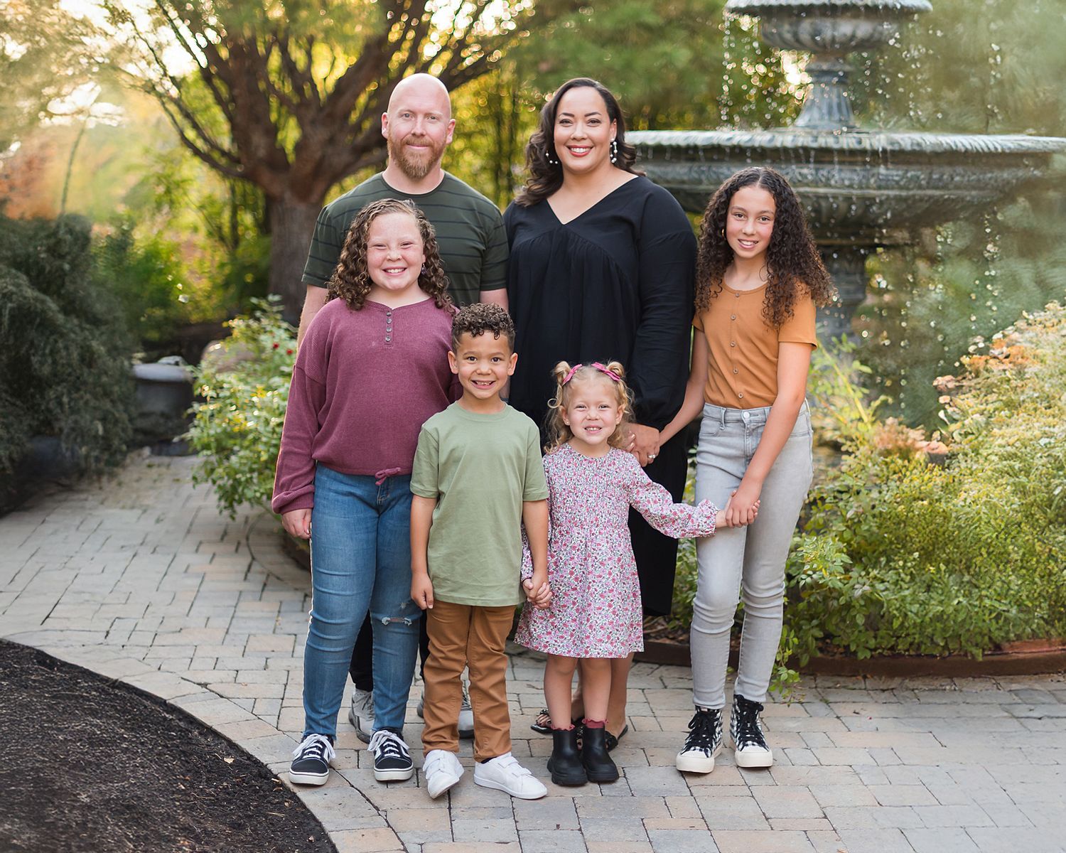 A happy family of five stands together on a garden path near a fountain during a sunny autumn afternoon.
