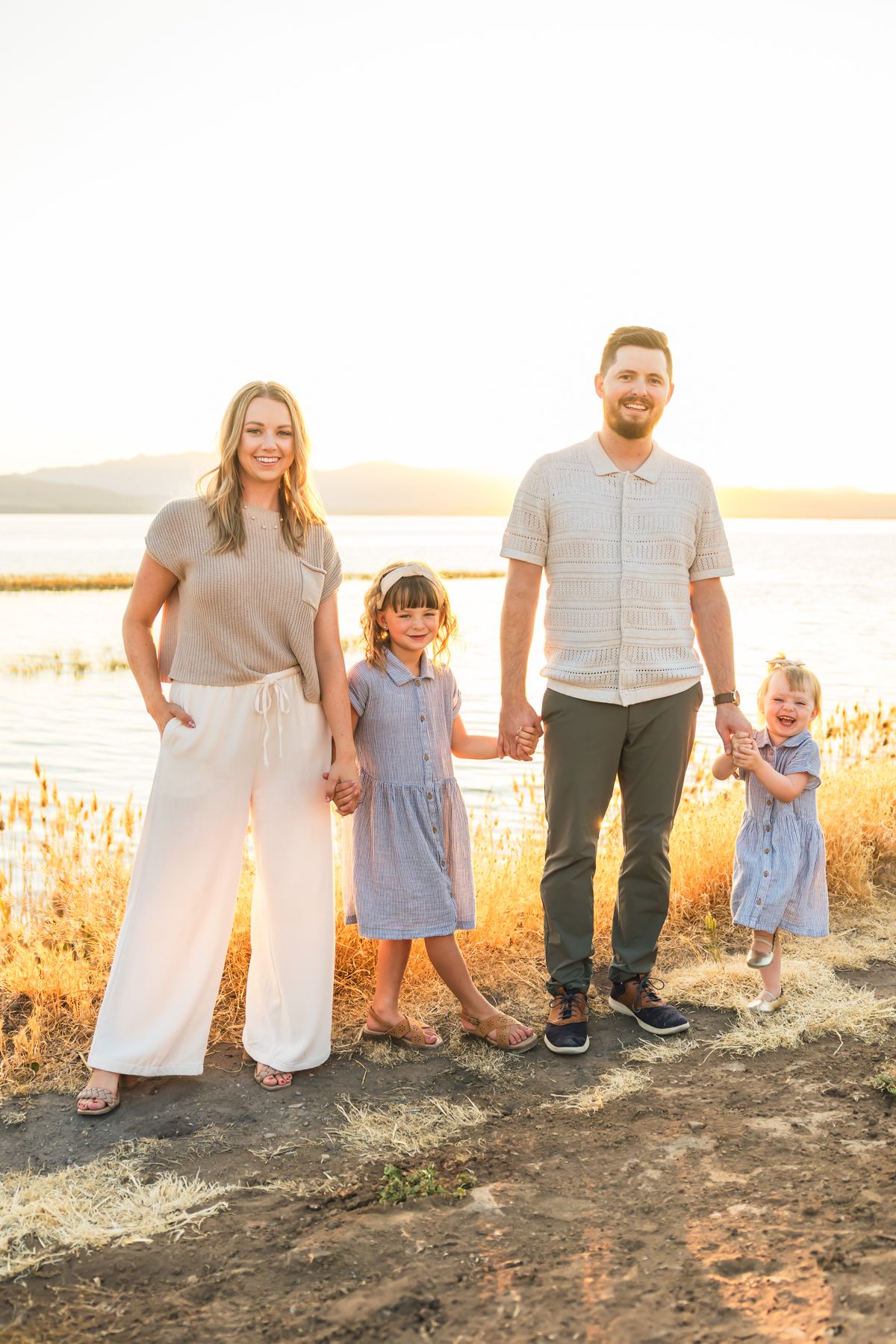 Family in coordinated outfits poses together at sunset by water with golden light and tall grass.