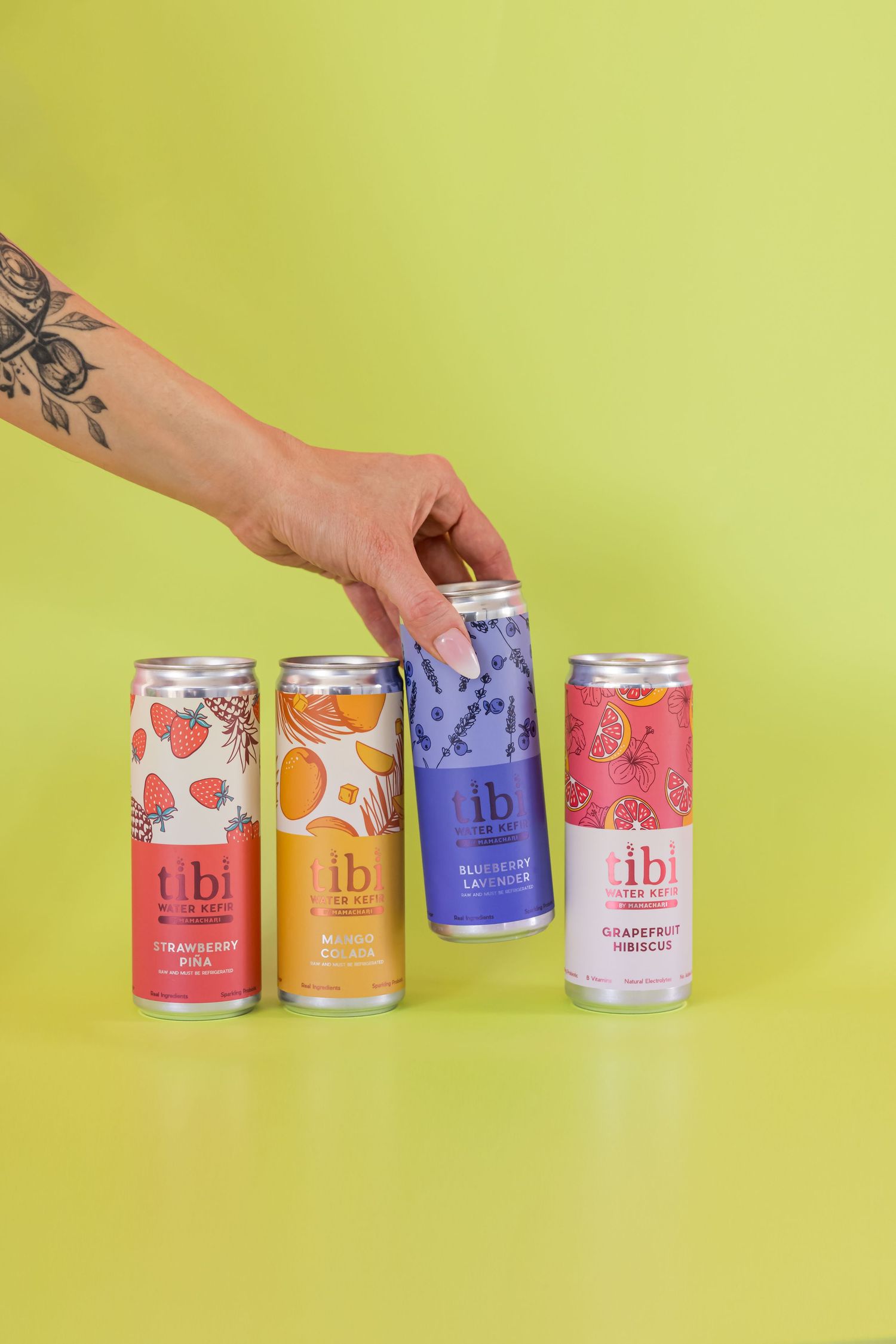 Four colorful cans of LB brand beverages lined up against a bright yellow background with a tattooed hand reaching.