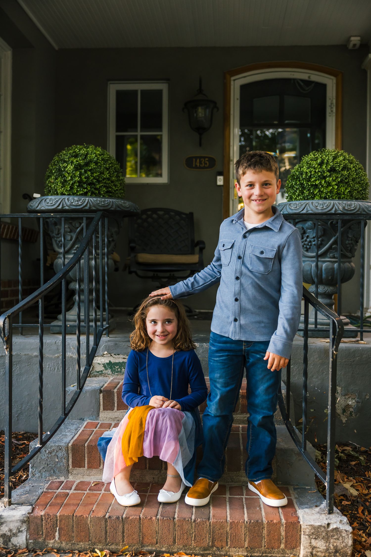 Two young siblings pose on their front porch steps in casual attire.