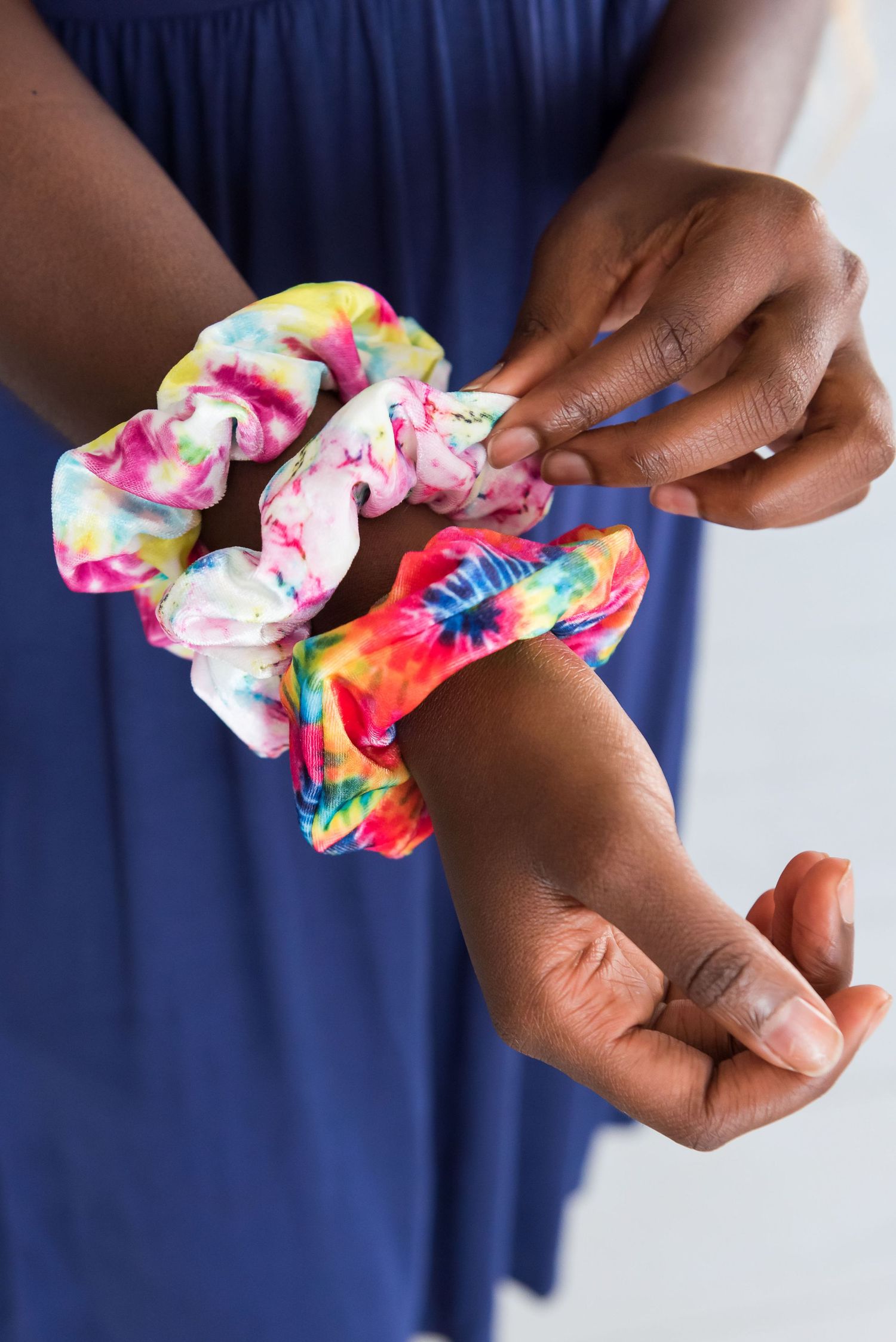 Hands wearing tie-dye scrunchie in bright rainbow colors against navy blue fabric.