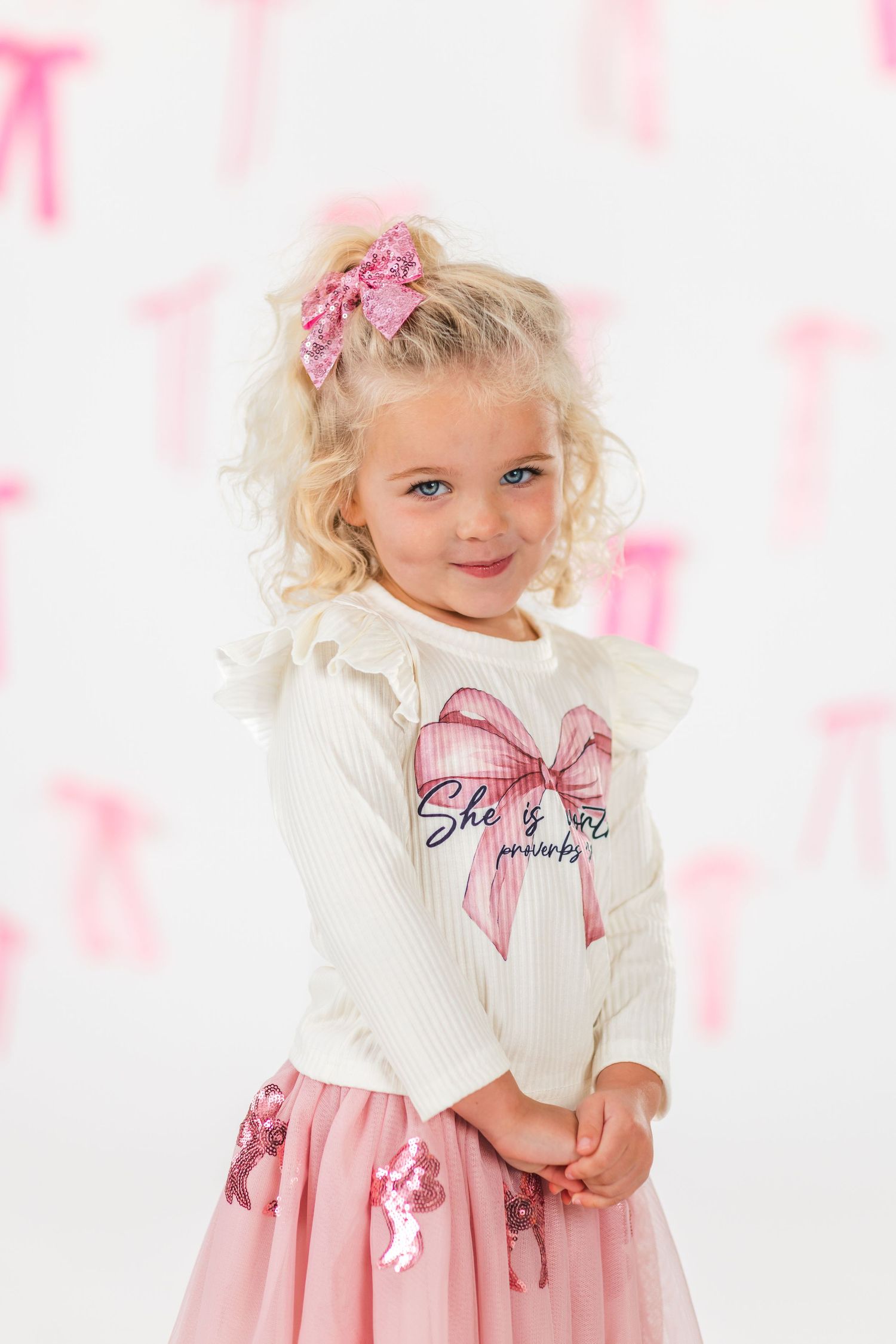 A child in a white sweater and pink tulle dress poses against a pink and white backdrop with floral accents.