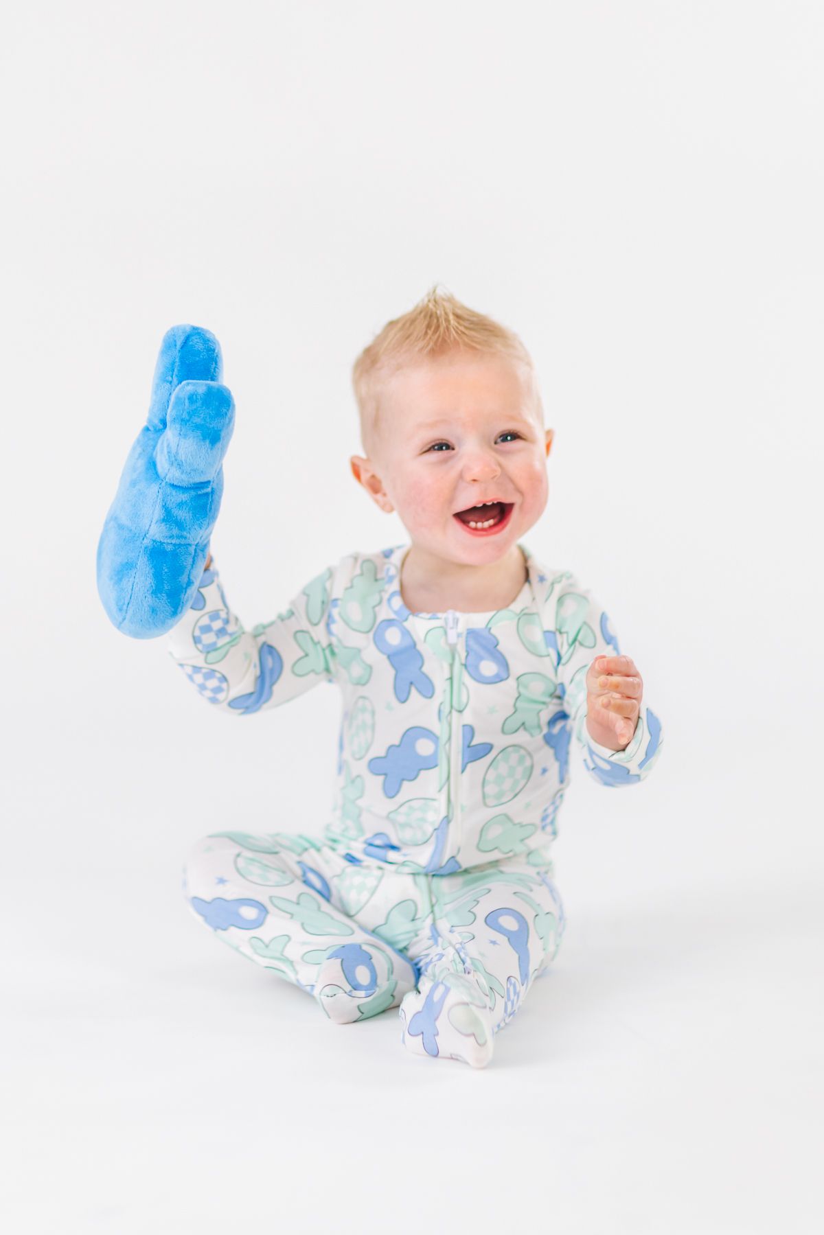 Laughing baby in blue and white patterned pajamas holding a blue stuffed toy on white background.