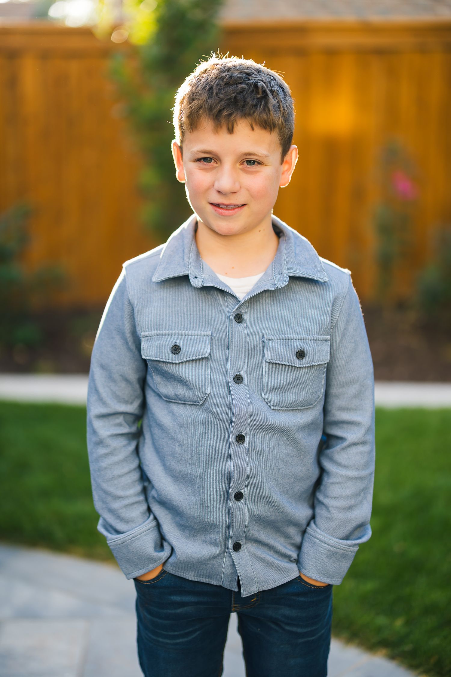Child in gray button-up shirt standing in backyard with wooden fence.