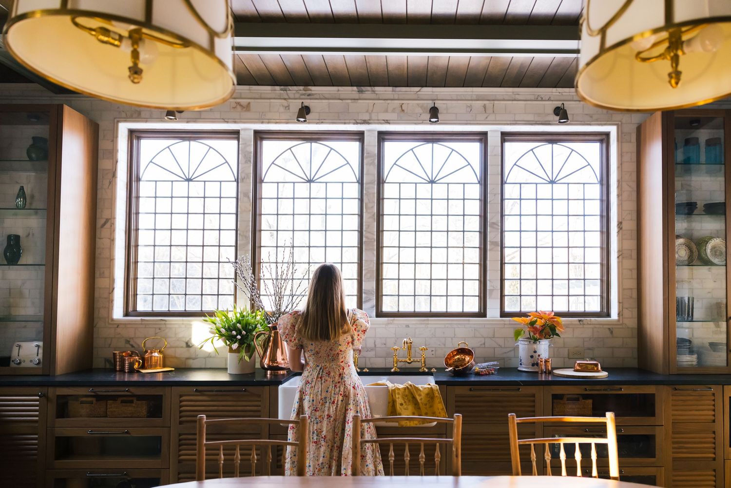 A rustic kitchen interior features arched windows, pendant lights, and wooden cabinetry in warm natural lighting.