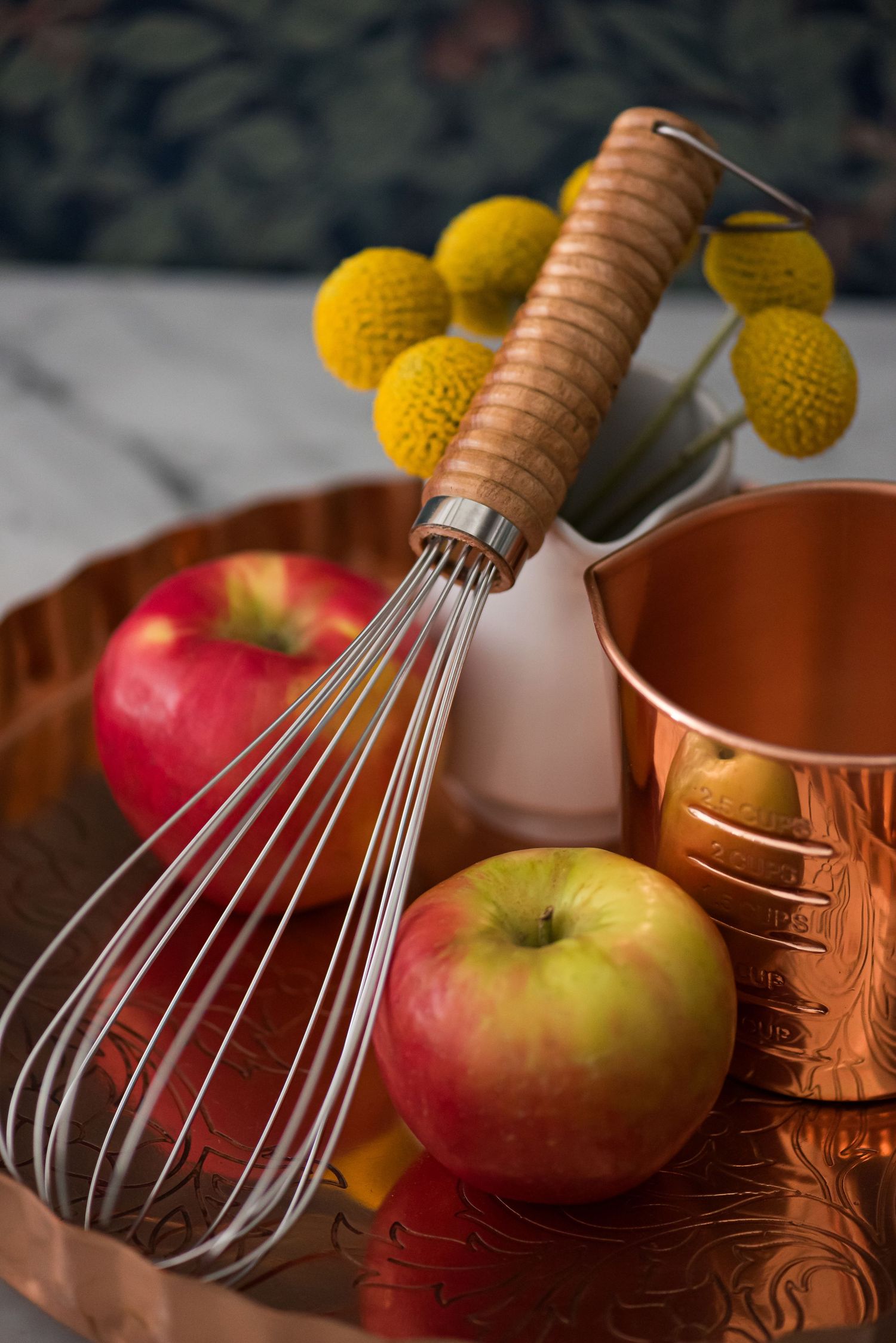 Copper whisk and apples with yellow floral accents in a rustic kitchen setting.