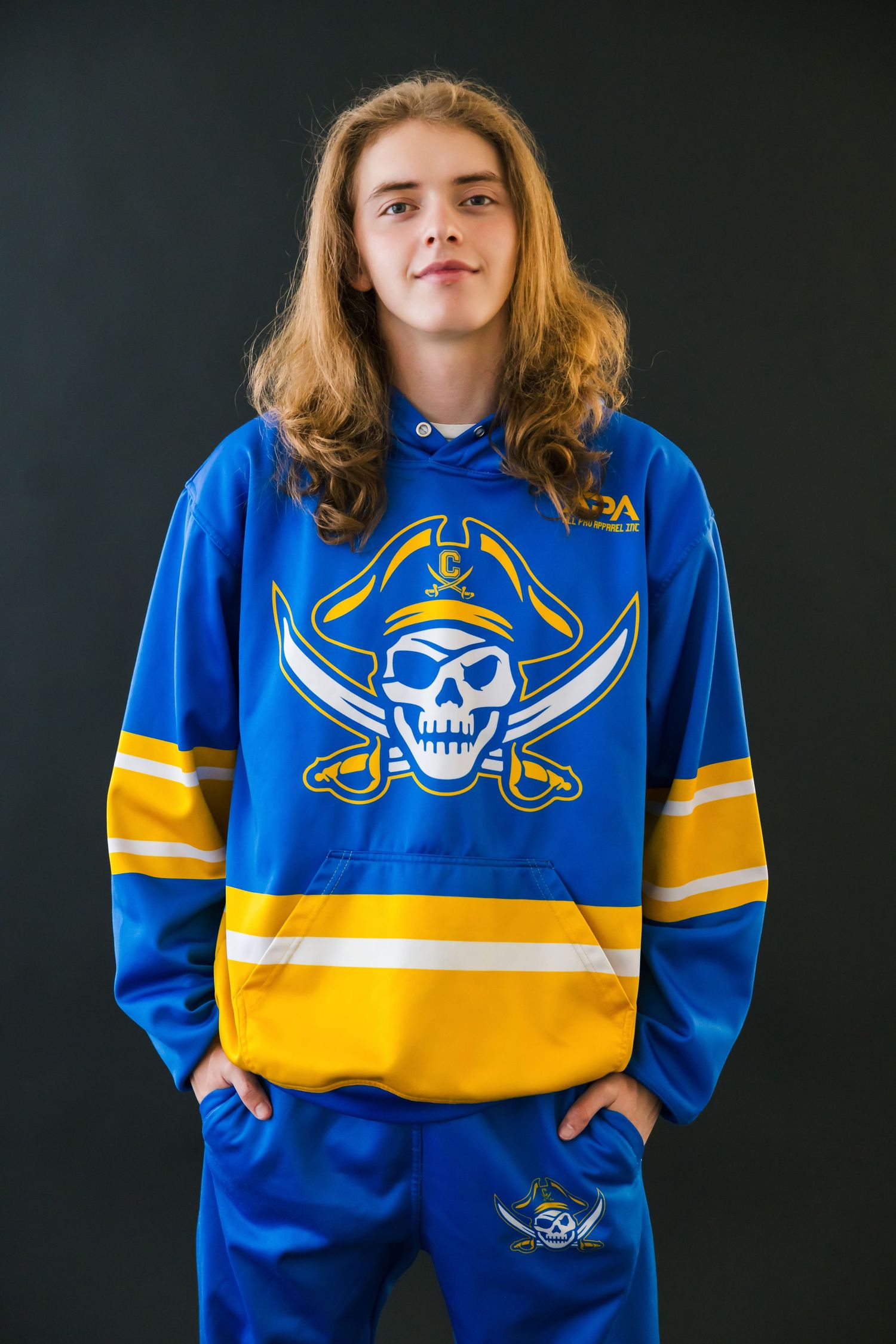 Young athlete in blue and gold pirate-themed hockey hoodie and matching pants poses against dark background.