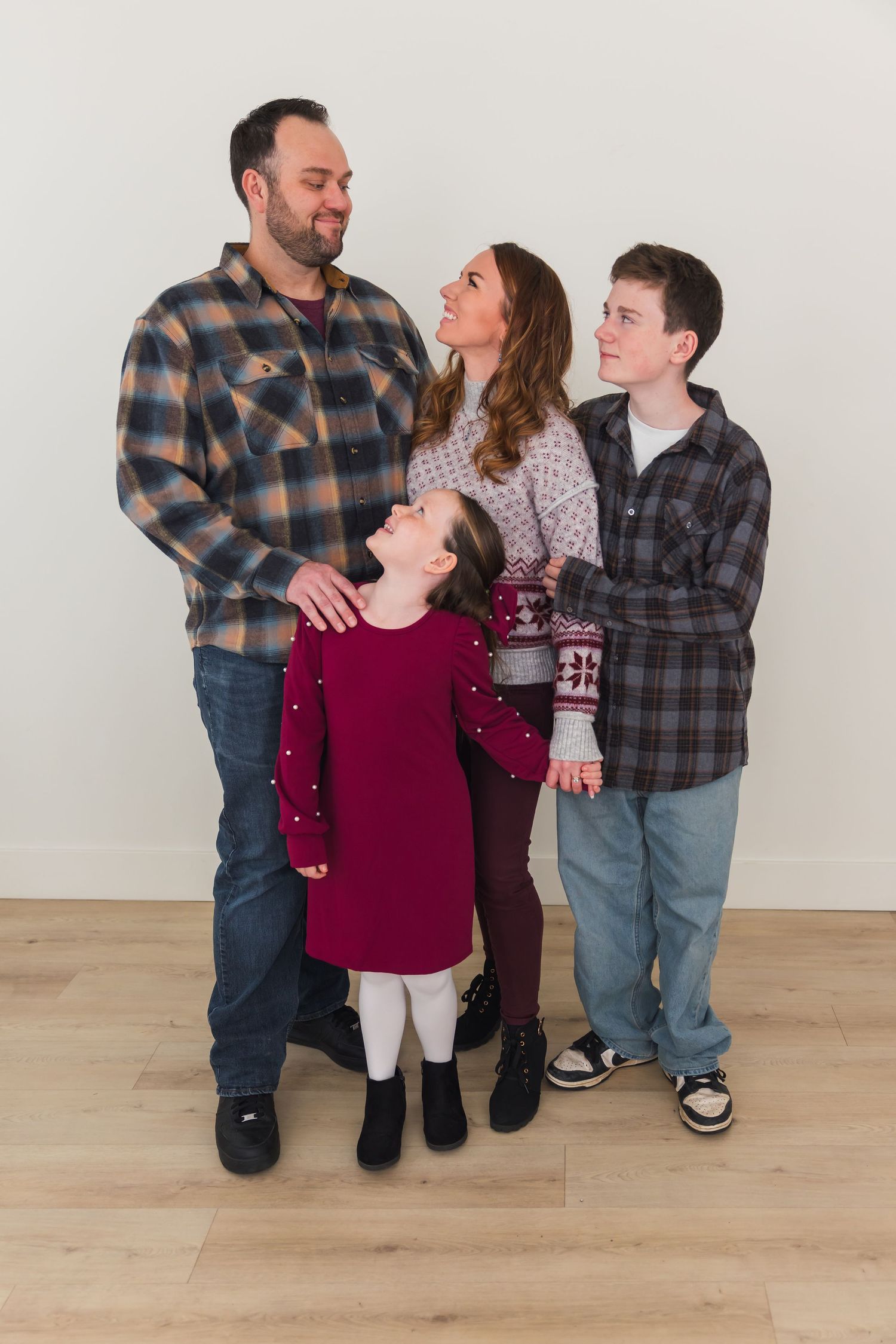 A family of four wearing plaid shirts and casual clothing poses together against a white wall on hardwood flooring.