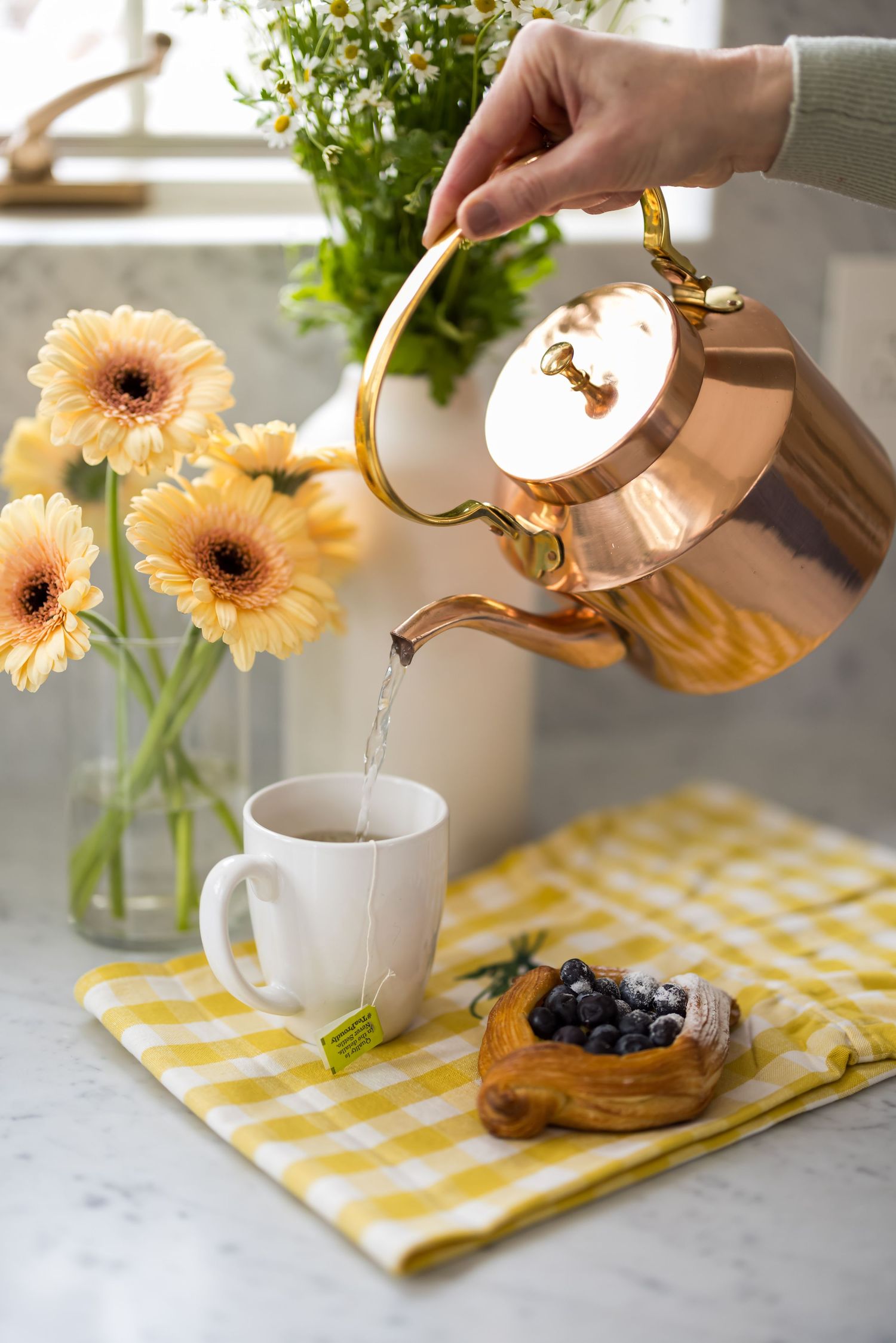 Tea being poured into white mugs on yellow checkered cloth with peach gerbera daisies and blueberries on table.