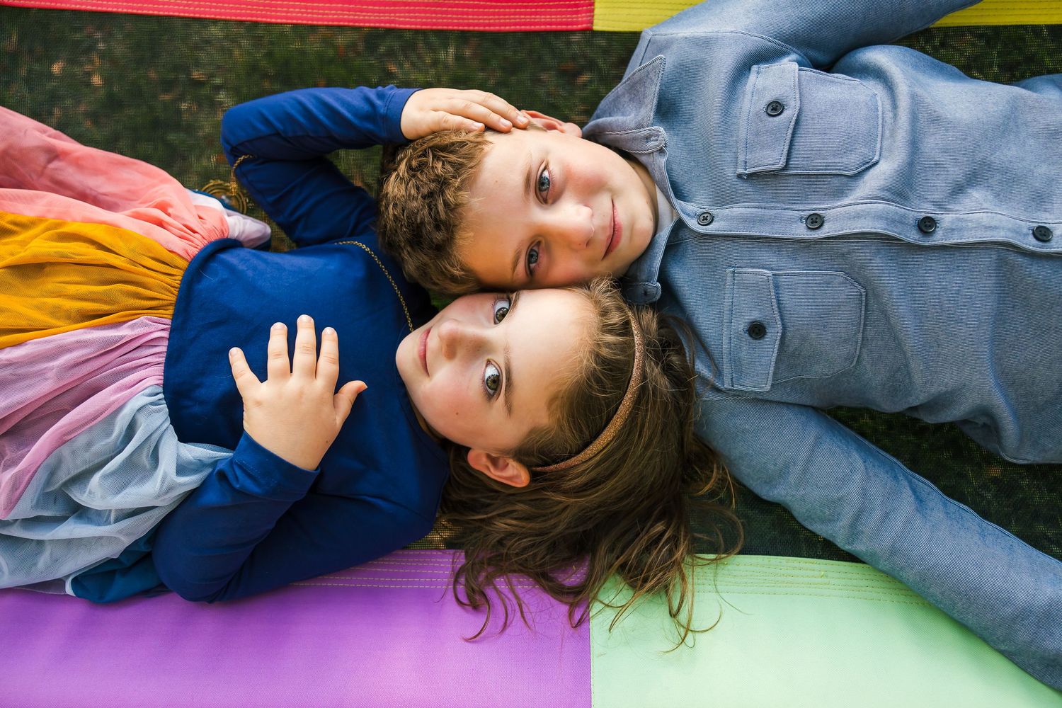 Two children lay on a colorful blanket outdoors while sharing a playful moment.