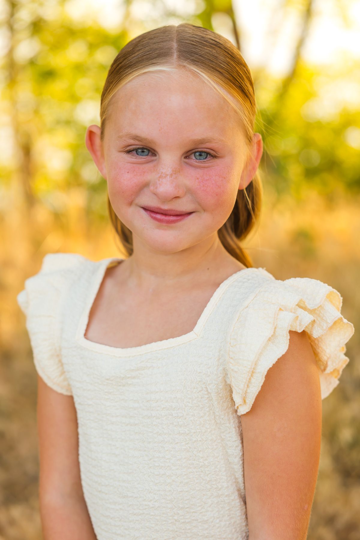 Portrait of a young person in a white textured top smiling against golden sunlit foliage.