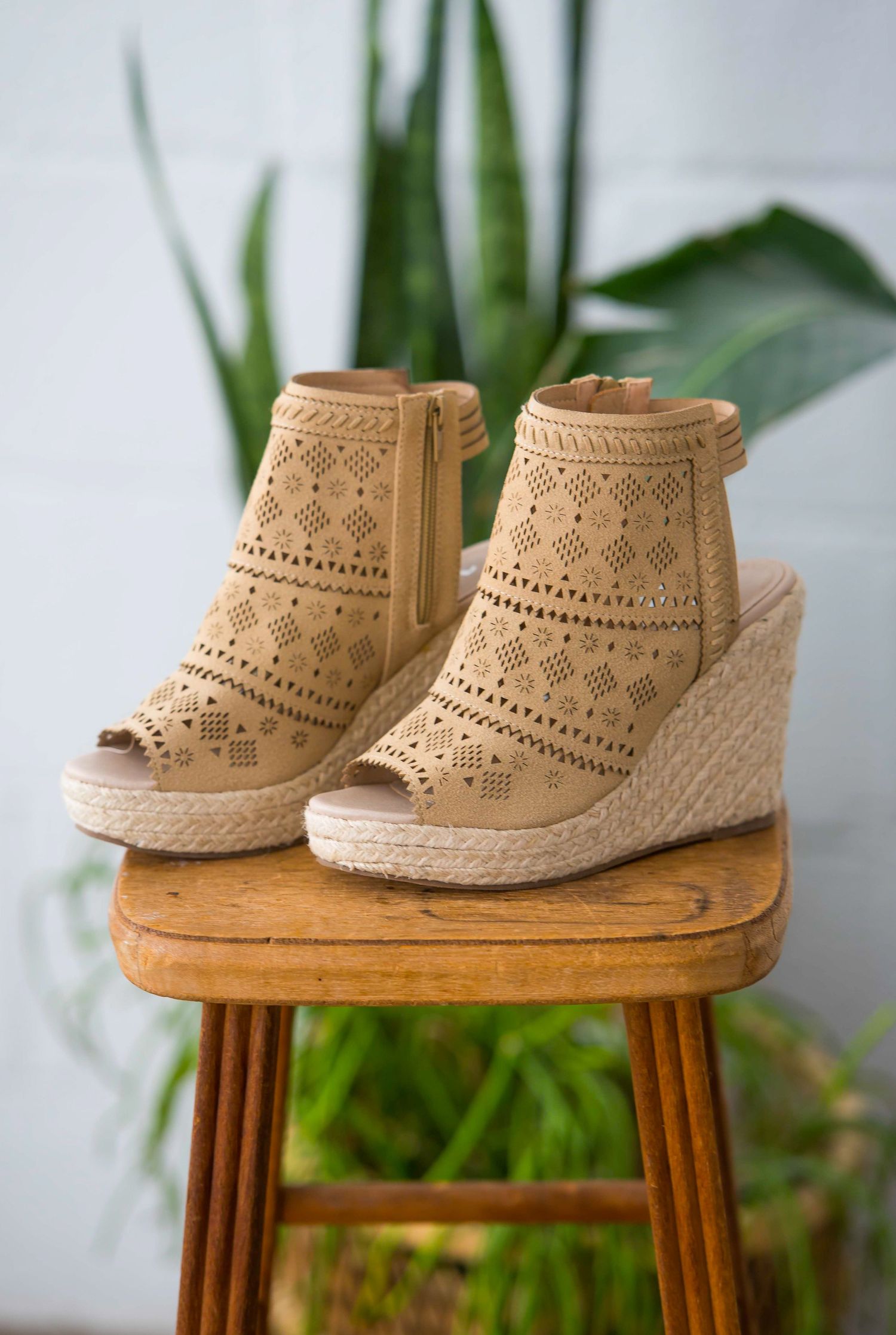 Beige crochet wedge sandals with cutout pattern displayed on wooden stool with plant background.