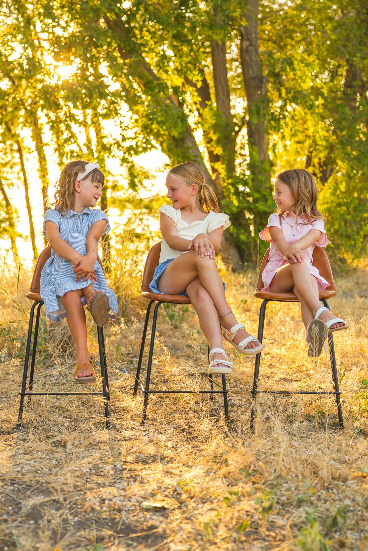 Three children sitting on tall chairs outdoors at sunset share laughs together in a golden-lit wooded setting.