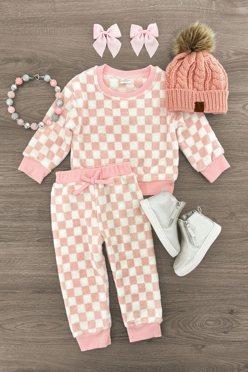 Pink and white checkered baby outfits with matching accessories and white shoes laid flat on wooden floor.
