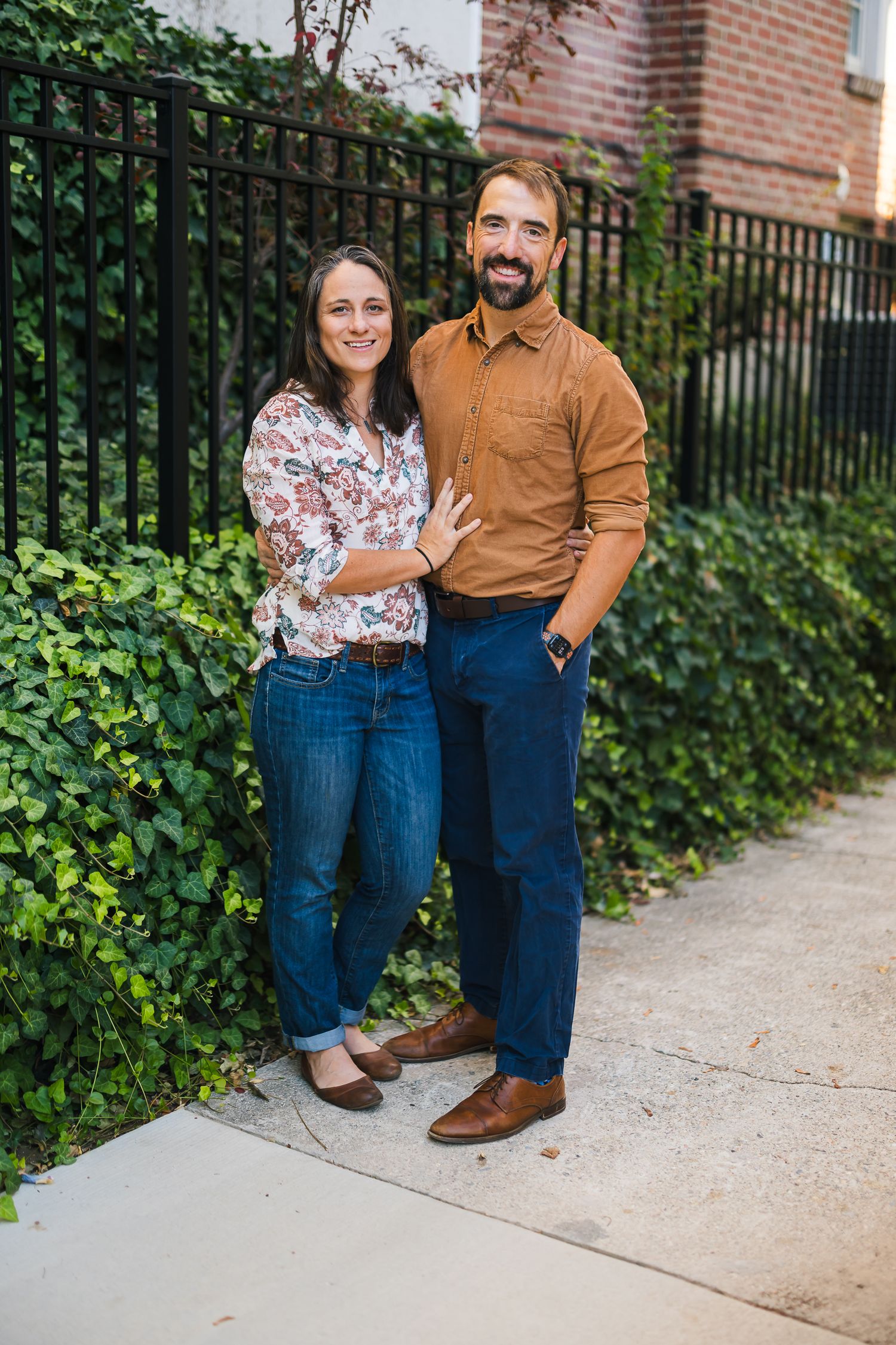 Outdoor portrait of couple in coordinating casual outfits against ivy-covered fence.