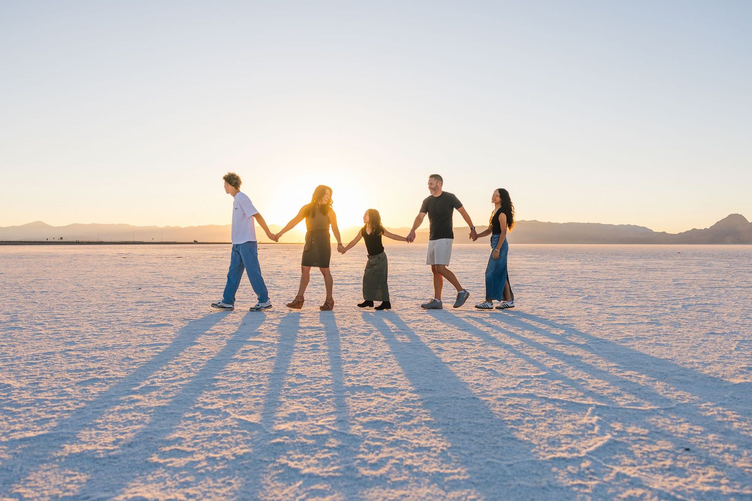 A group walking hand in hand across a pristine salt flat at sunset casting long shadows on the white ground.