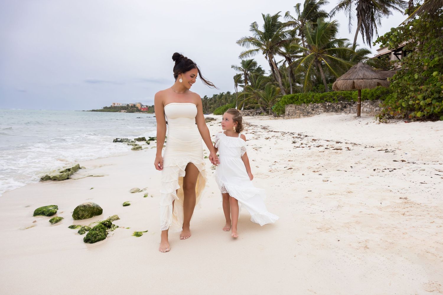Two people in white dresses walking together on a tropical beach with palm trees.