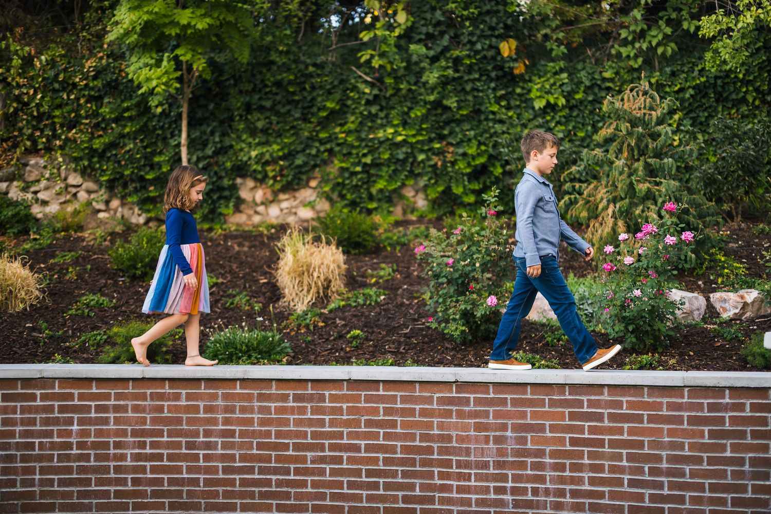 Two people walking in opposite directions along top of brick retaining wall in garden setting.