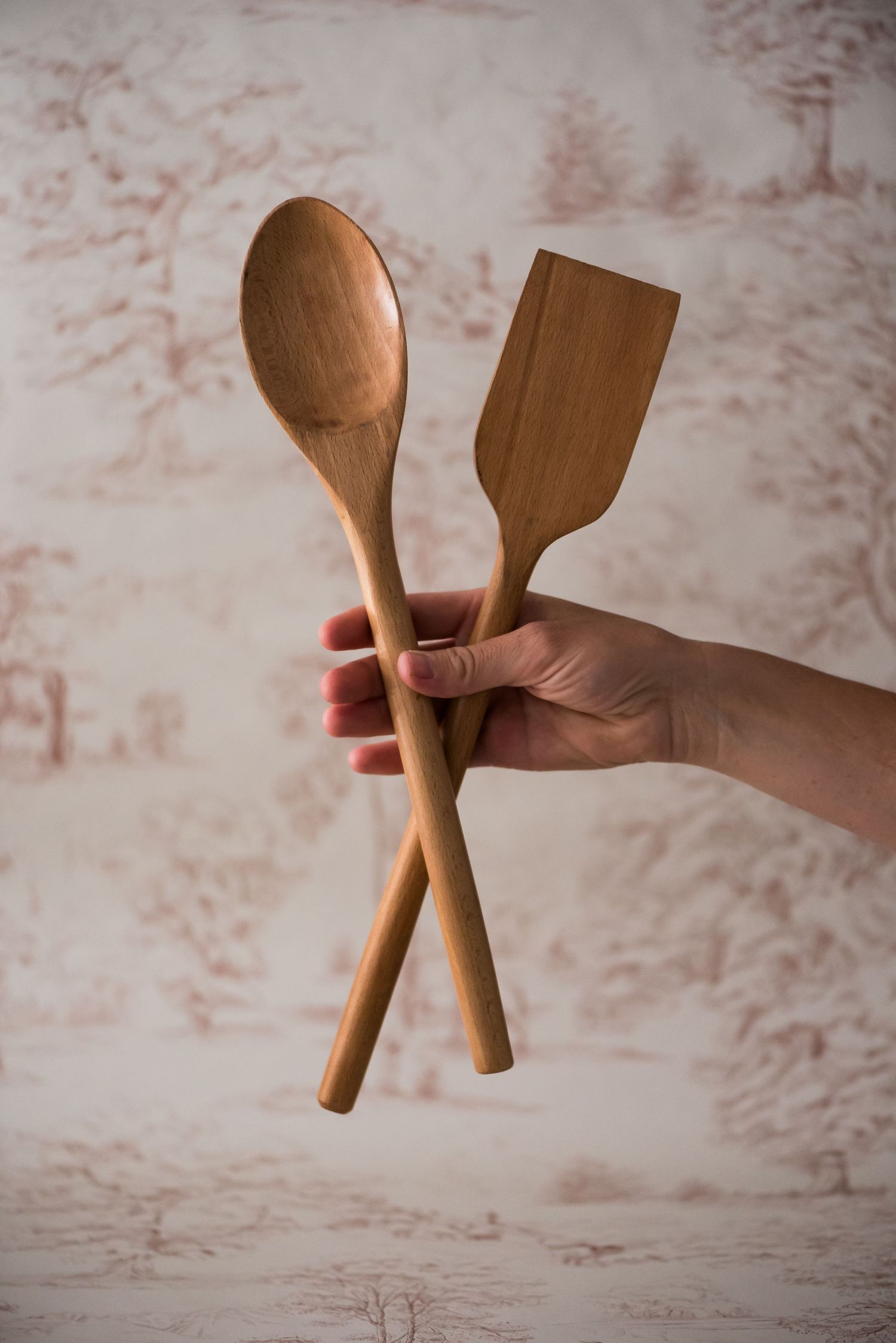 Hand holding crossed wooden cooking utensils including a spoon and spatula against textured beige background.