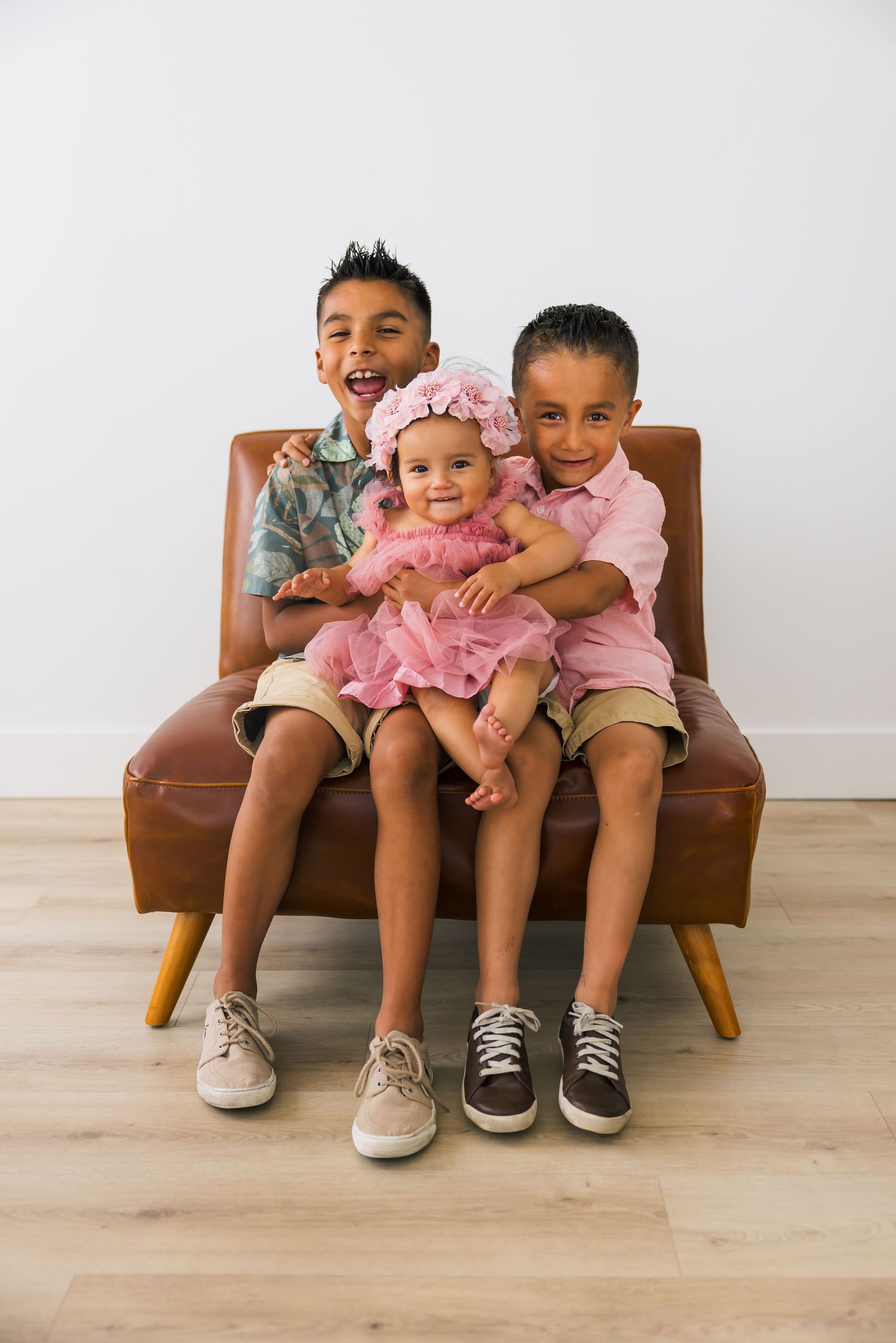Two children sitting together on a brown leather bench against a white wall in three similar poses.