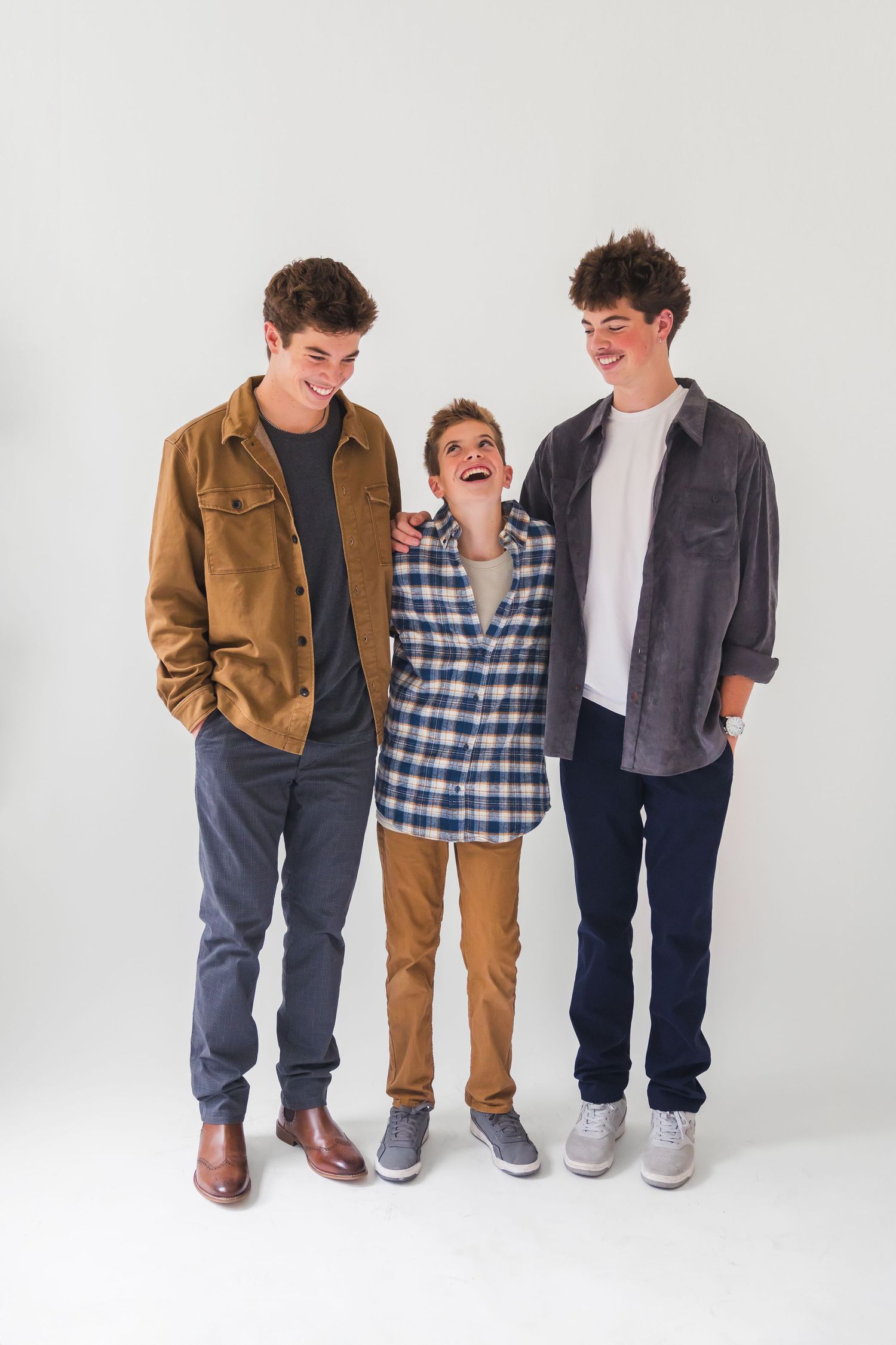 Three children stand together in casual clothing including plaid shirts, jeans and brown jackets against a white background.