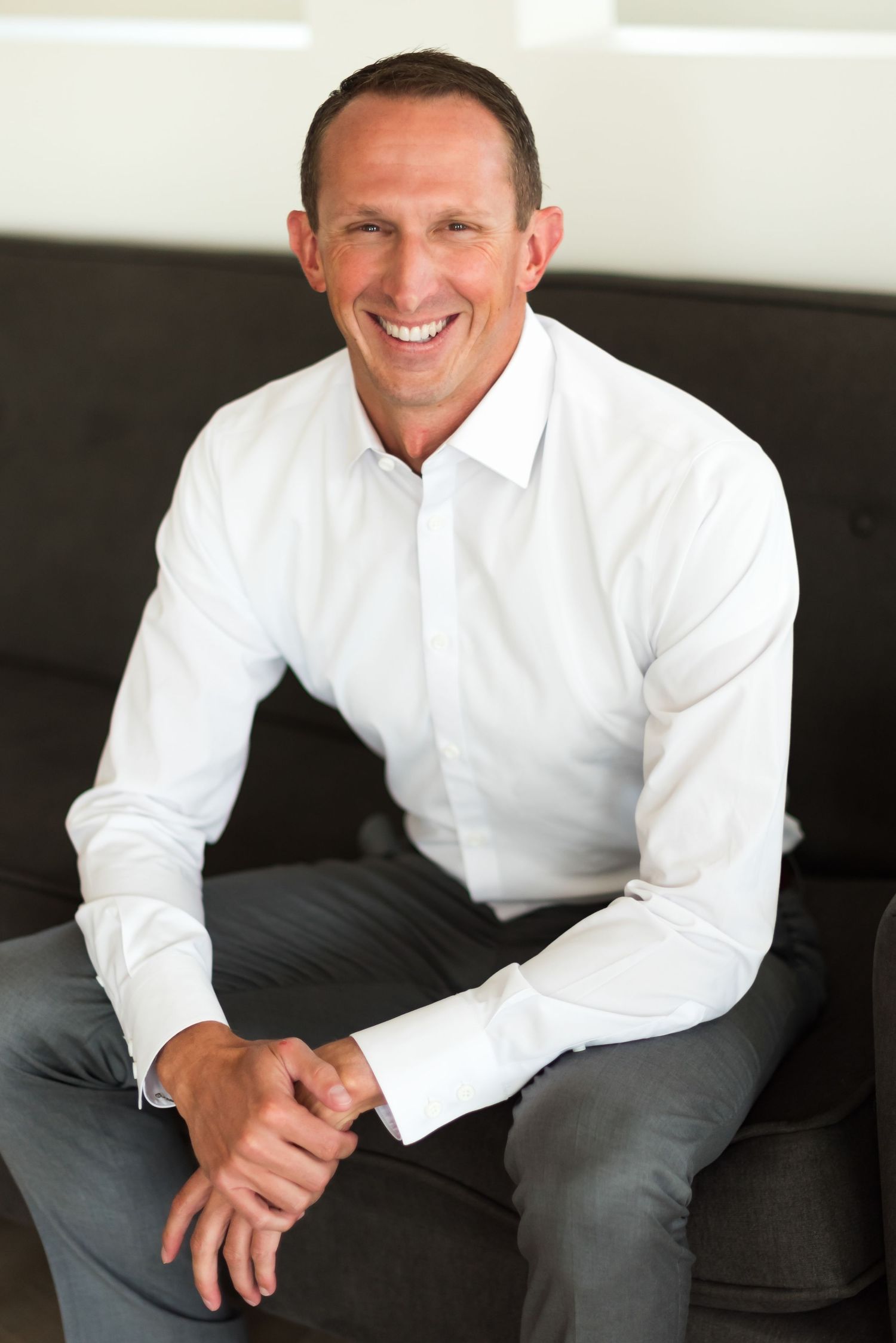 Professional portrait of a person in white shirt and gray pants seated on dark couch.