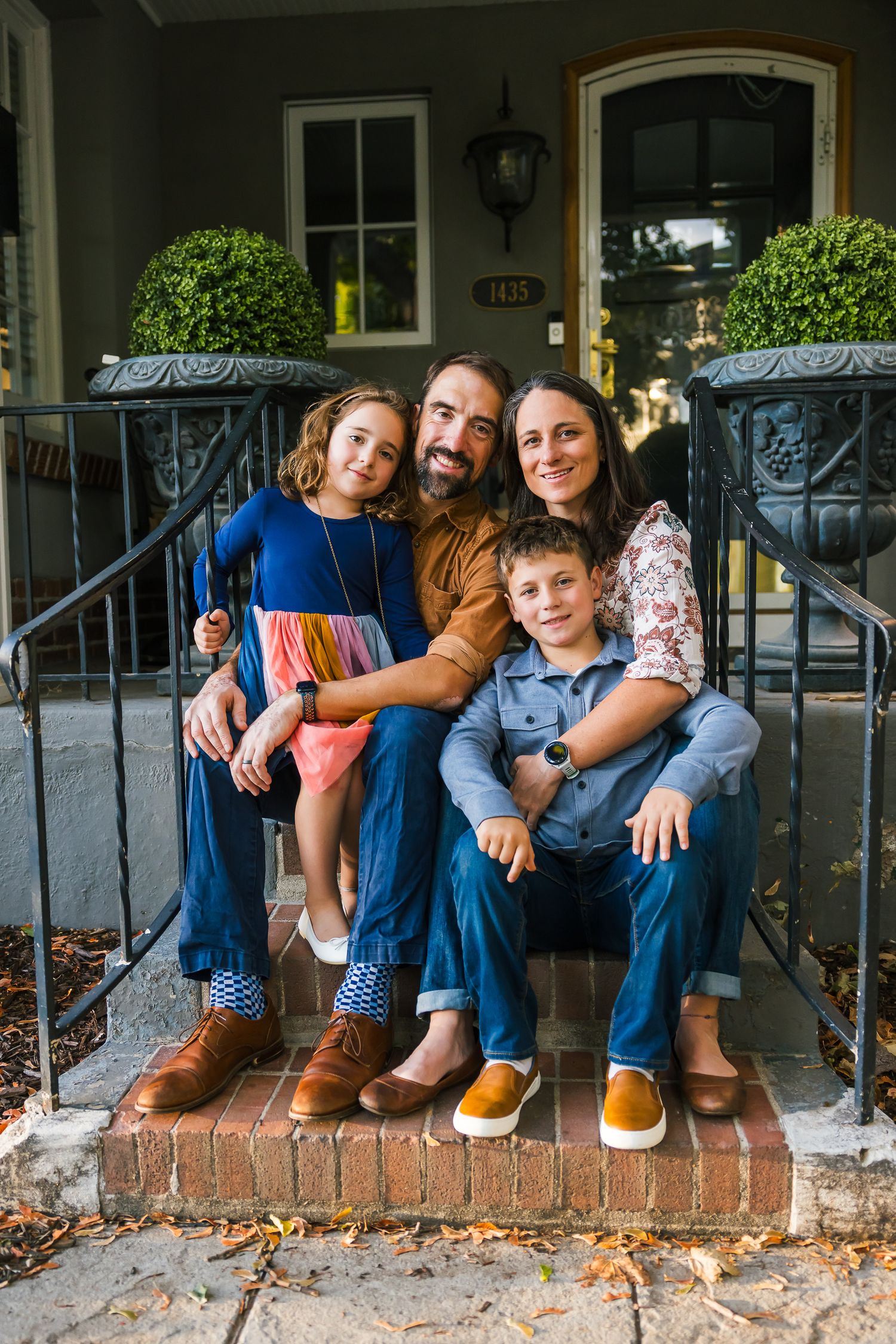 A family of four sits together on brick front porch steps with decorative railings.