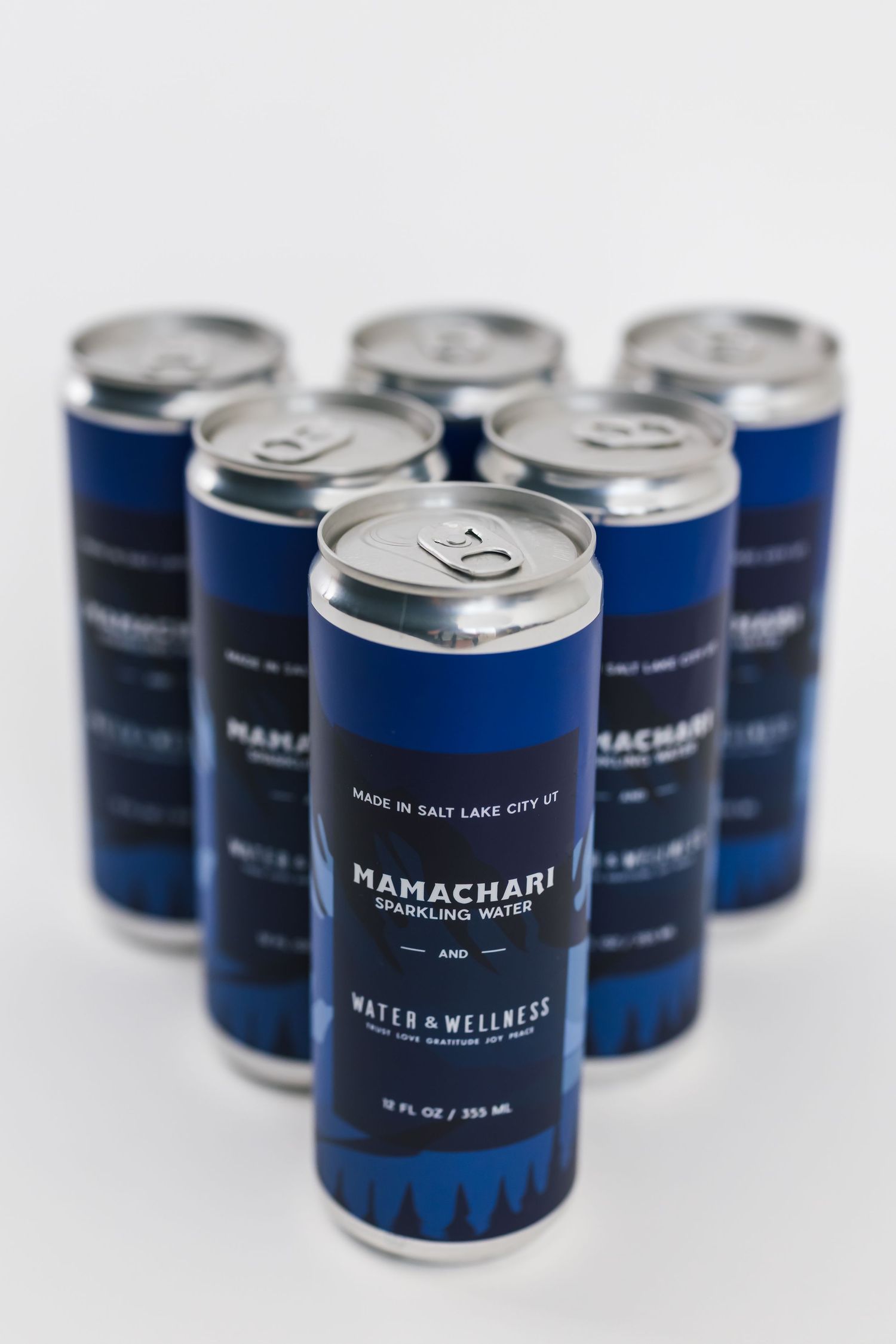 Six blue aluminum cans of Mamachari water and wellness drinks arranged in a triangular formation on white background.