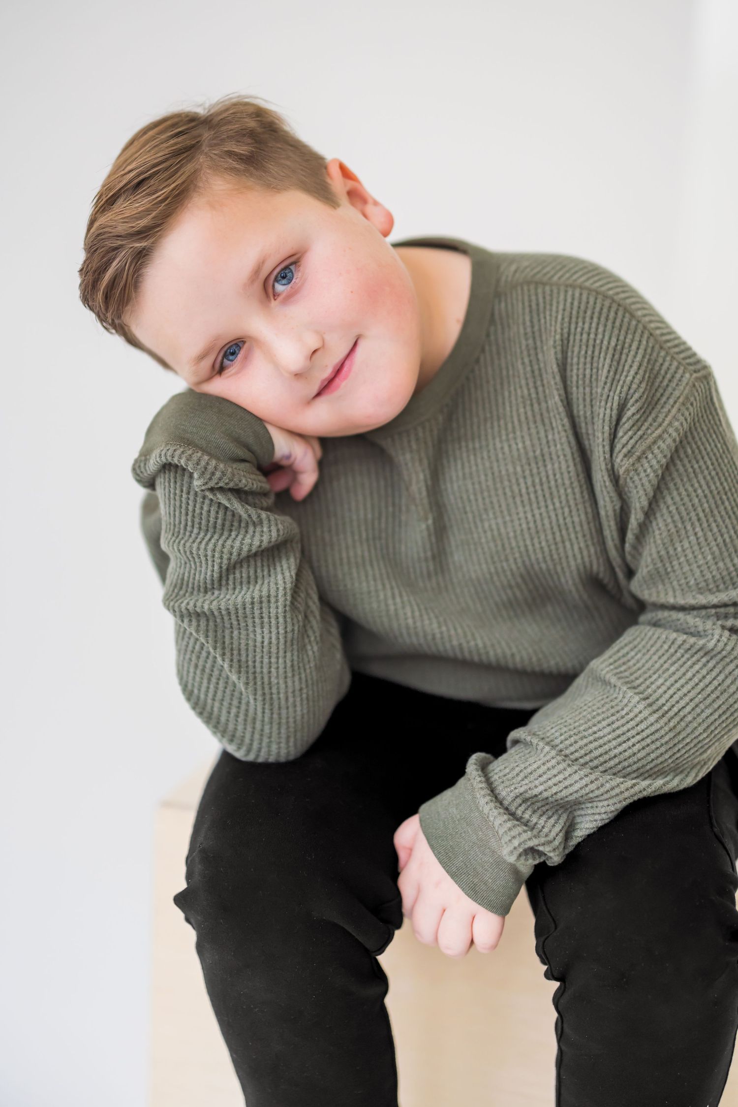 A young person wearing a sage green sweater and black pants poses playfully against a white background.