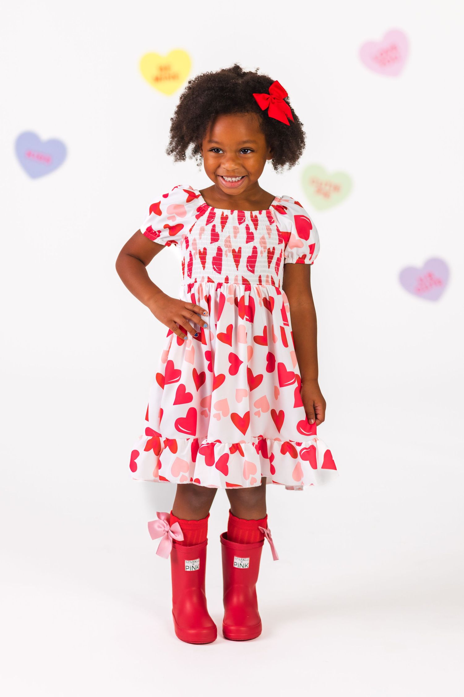 A child wearing a white and red heart-patterned dress with red boots poses against a white background with heart decorations.