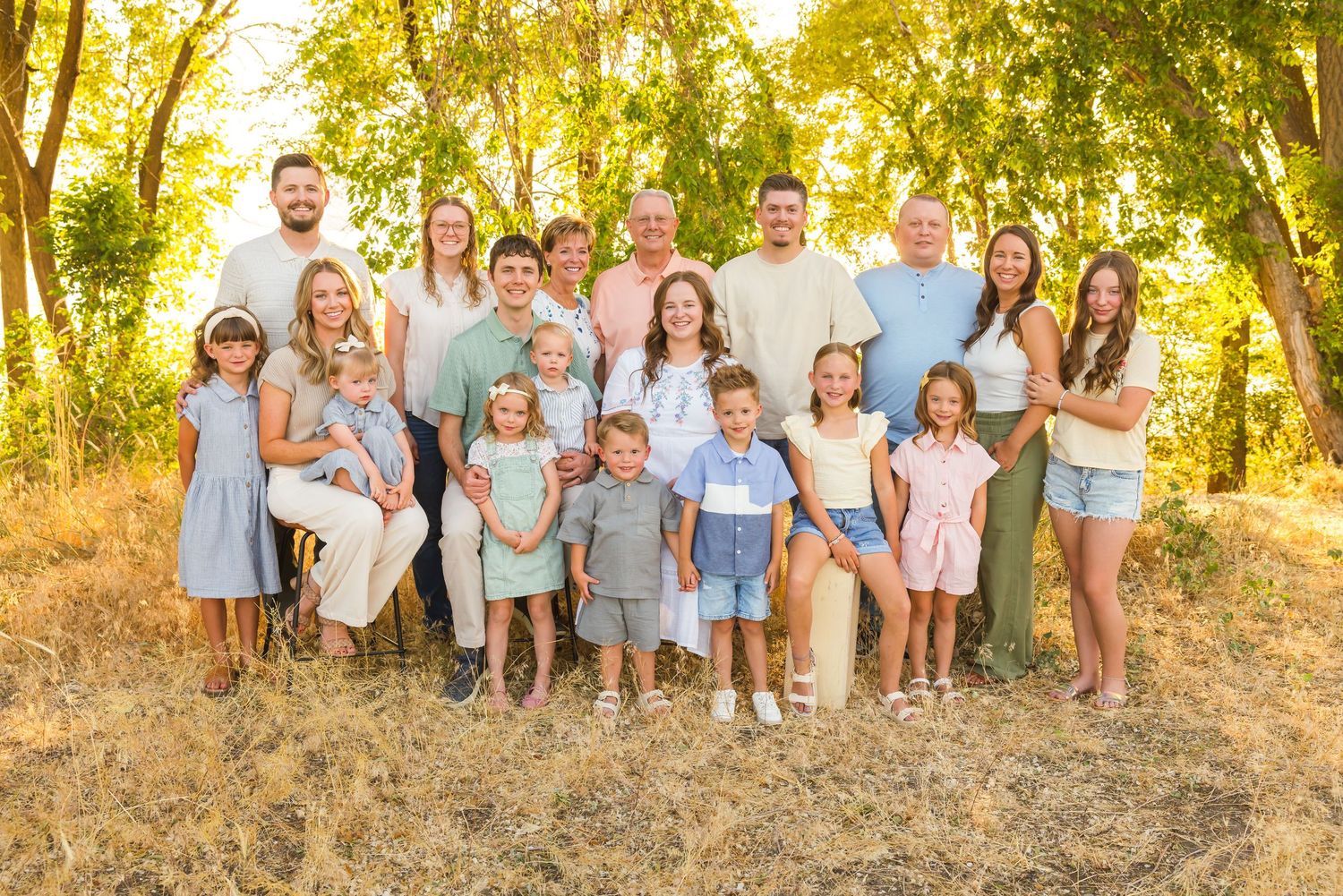 A large family group in coordinating blue and neutral outfits poses together at sunset in a golden wooded area.