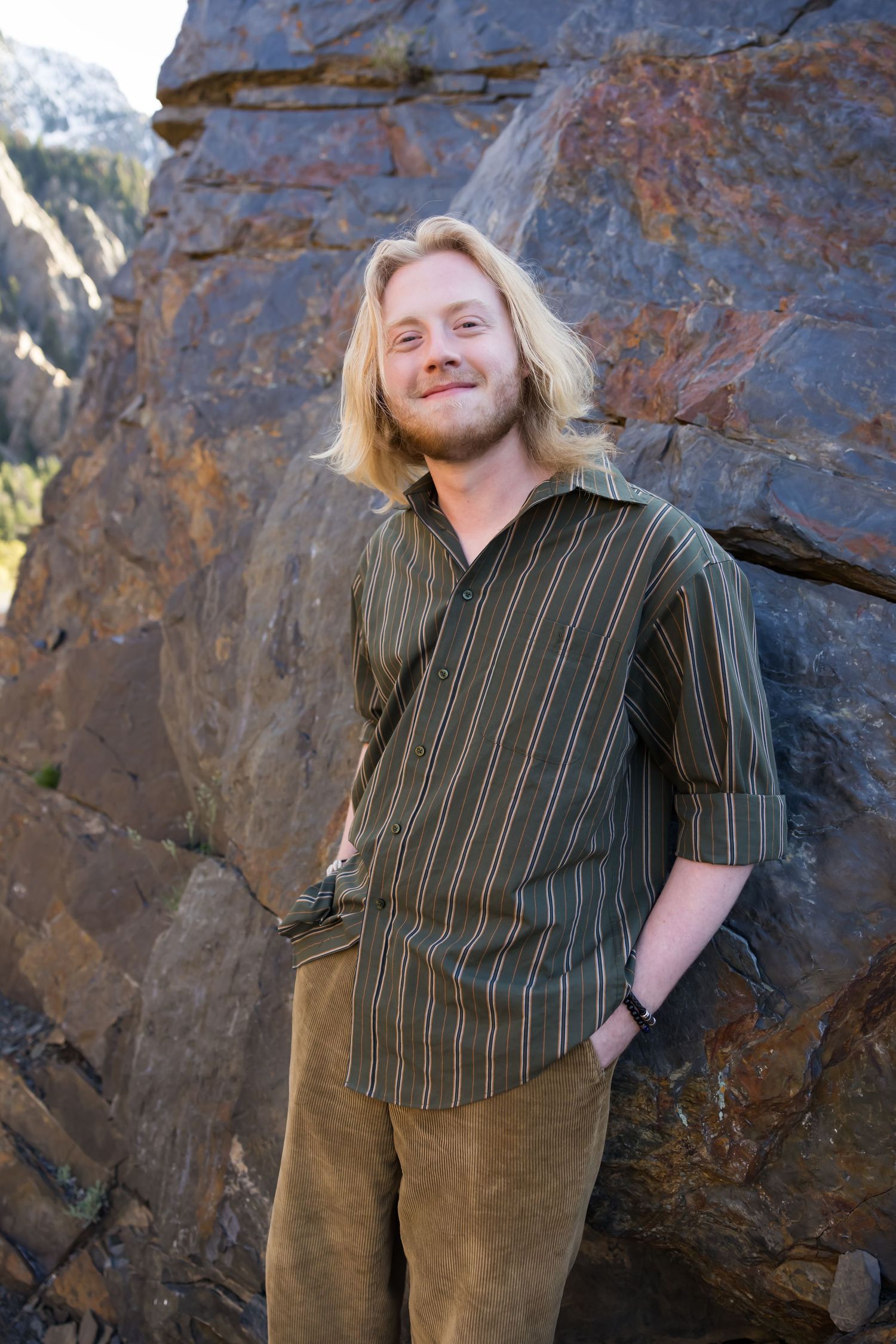 Person in striped button-down shirt stands against rocky cliff face in outdoor mountain setting during golden hour.
