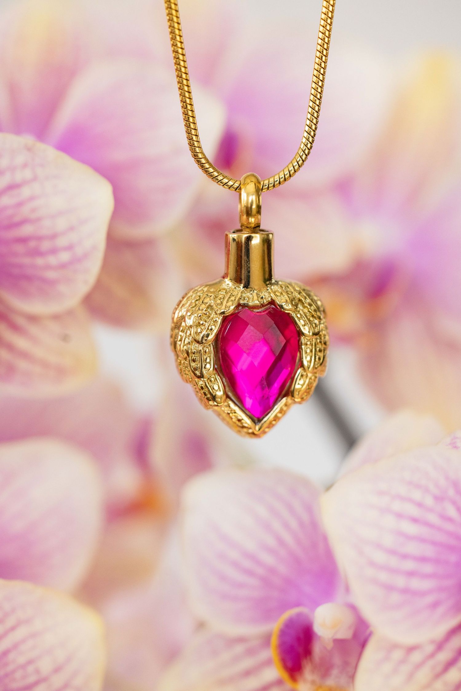 Gold heart pendant with pink gemstone on chain against blurred pink floral background.