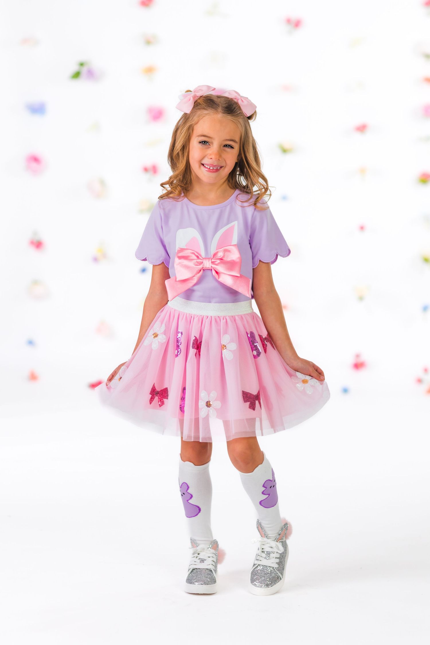 Young child wearing pink and purple dress with bow detail twirls and poses against white background with colorful confetti.