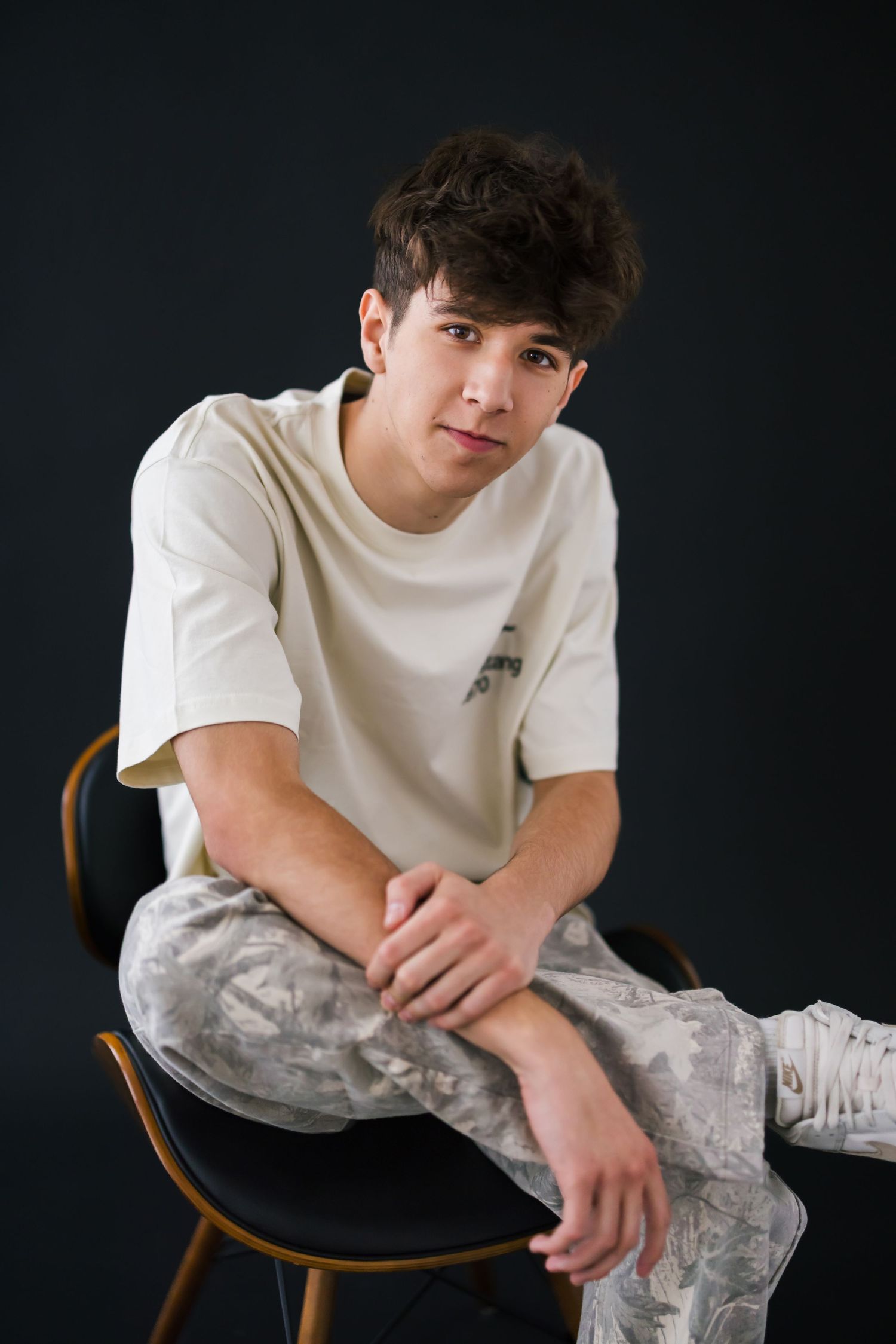 Young person in white t-shirt and camo pants seated on wooden chair against black backdrop.