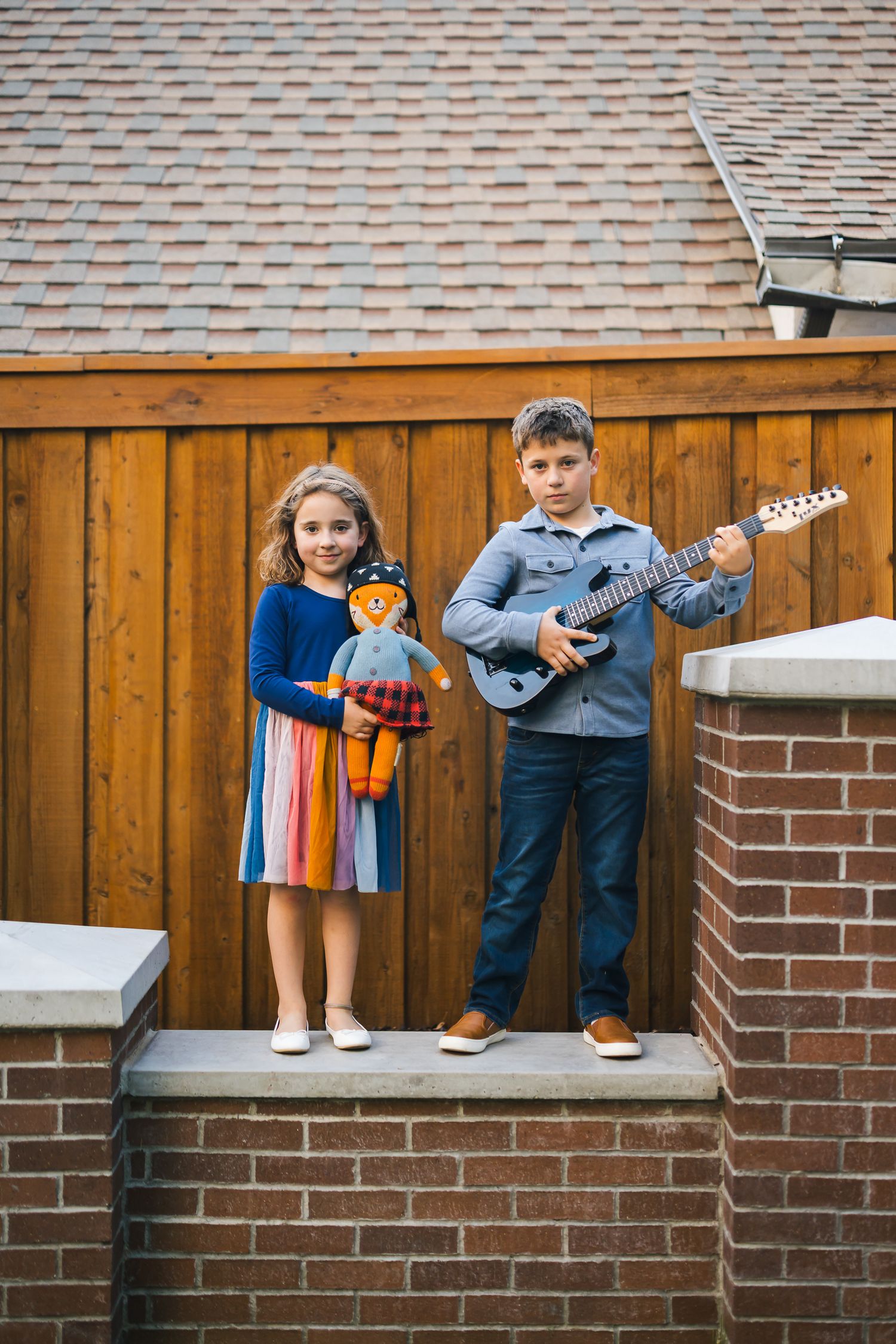 Two children stand on brick steps, one holding a puppet and one with electric guitar.