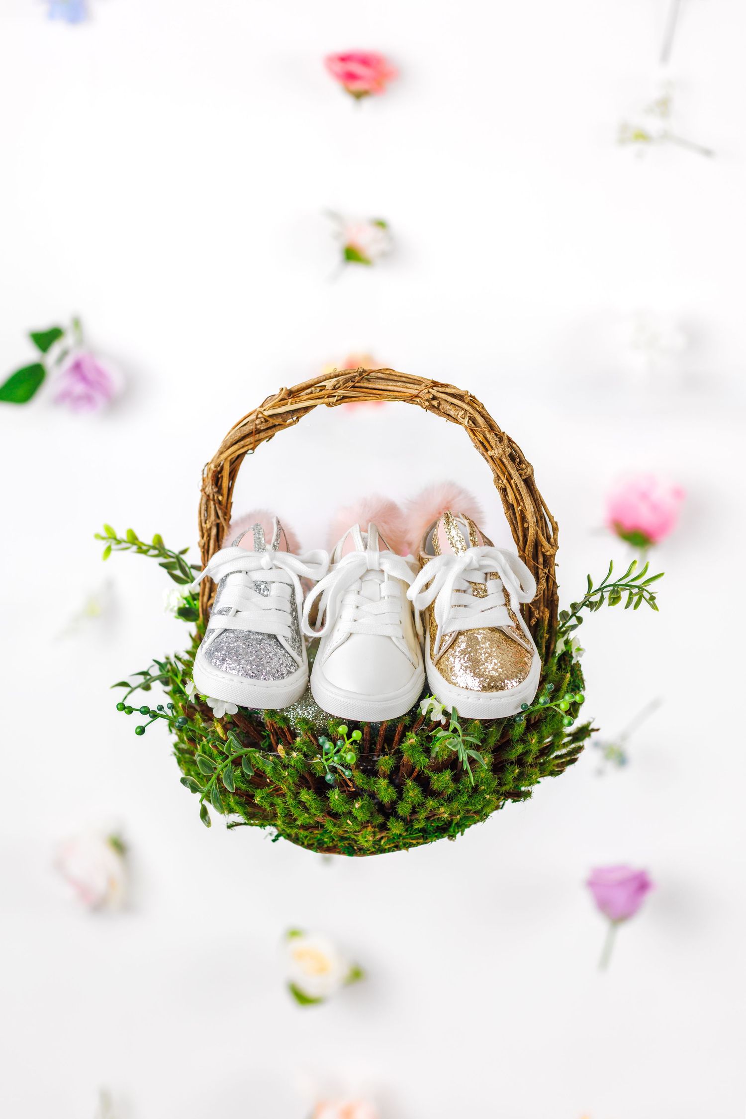 Tiny baby shoes nestled in moss-filled wicker baskets with delicate pink flower petals scattered around.
